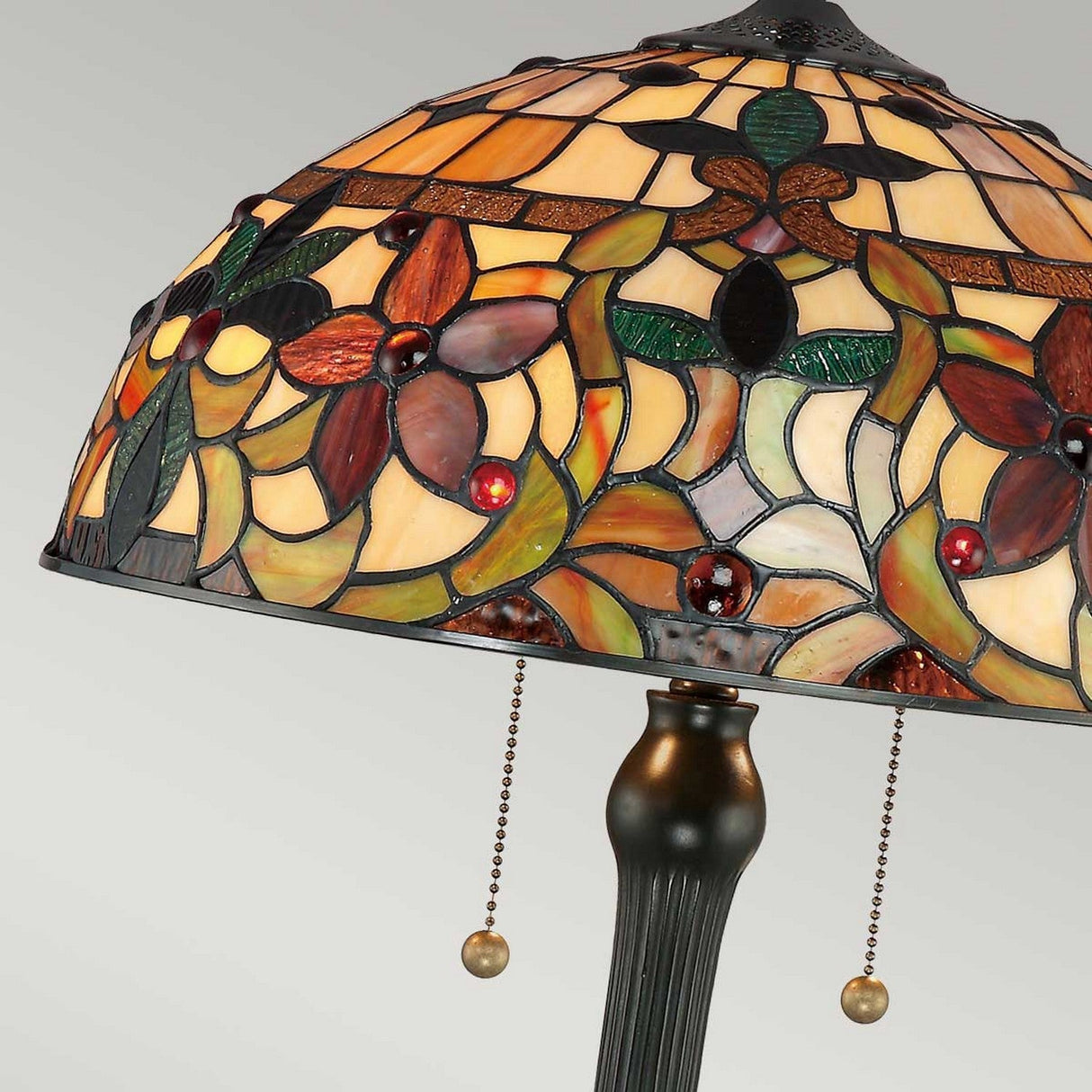 The Kami 2 Light Tiffany Table Lamp in Vintage Bronze features an art glass shade decorated with floral and geometric patterns in earthy tones. Its vintage bronze base is complemented by a dark, ribbed stand with two pull chains, adding to its timeless charm.