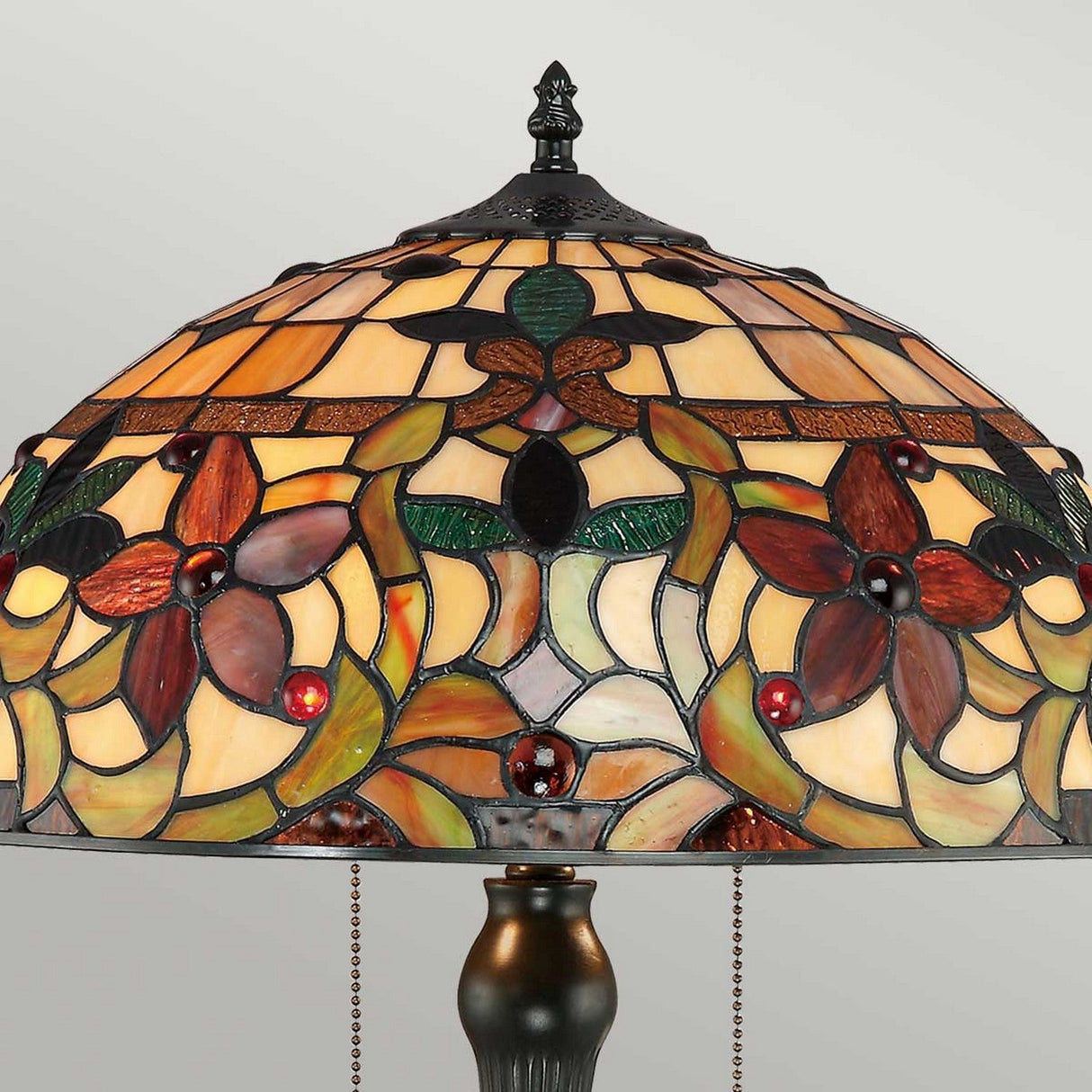 The Kami 2 Light Tiffany Table Lamp in Vintage Bronze boasts a decorative floral pattern with an elegant mix of amber, green, and red hues. Its vintage bronze base beautifully complements the intricate flower designs on the art glass shade, resulting in an elegantly artistic appearance.