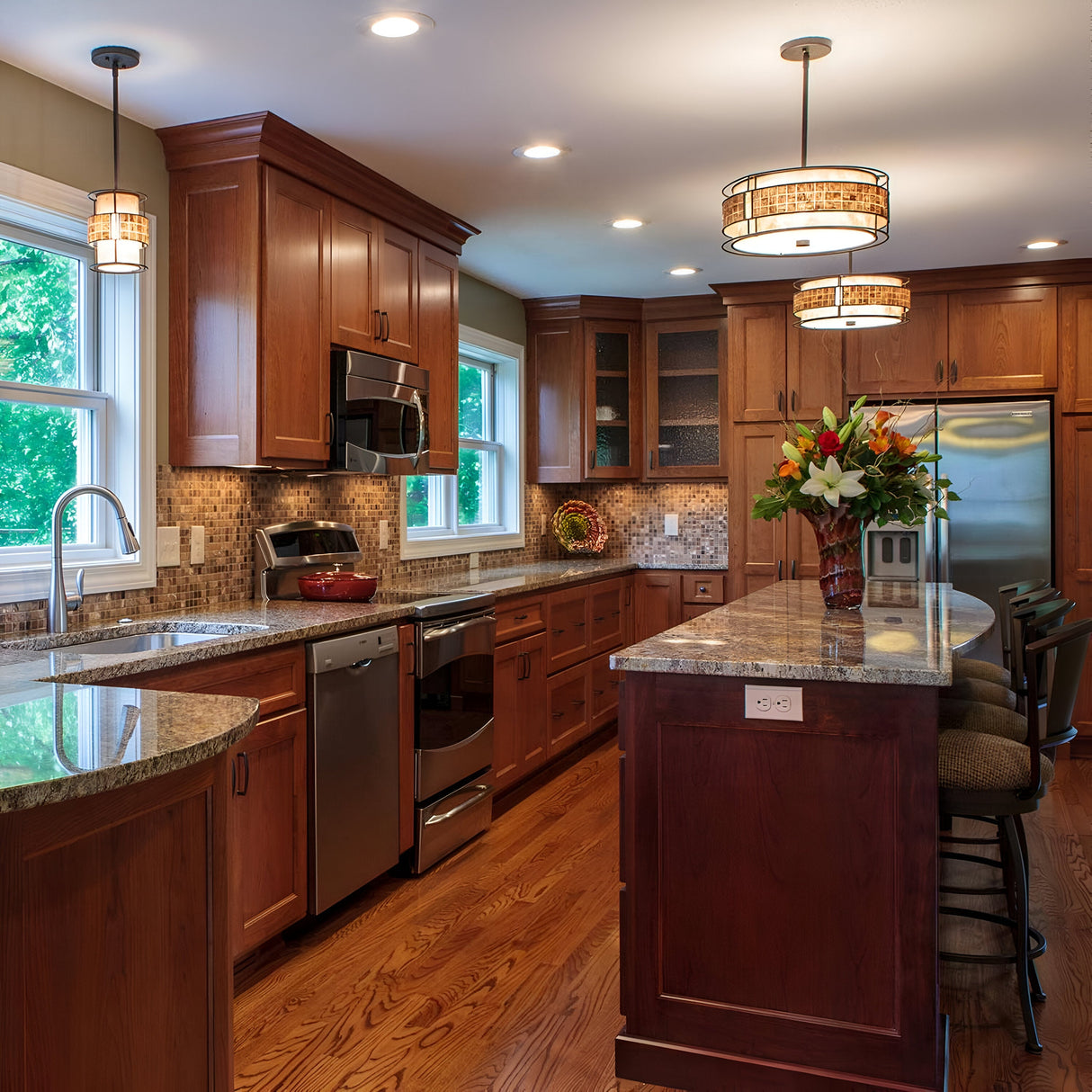 A spacious kitchen with wooden cabinets and a striking mosaic tile stripe backsplash complements the stainless steel appliances. The island features high-back chairs, a vase of flowers, and is illuminated by elegant Laguna 1 Light Mini Pendant fixtures in Renaissance Copper. Natural light floods in through two large windows.