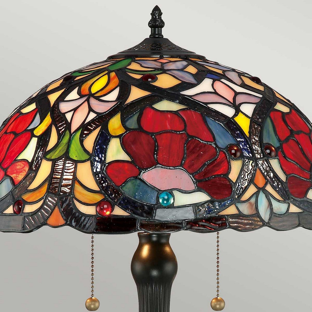 The Larissa 2 Light Tiffany Table Lamp in Vintage Bronze displays a detailed floral motif, highlighting the craftsmanship of handcrafted art glass. The design includes red, pink, yellow, and blue pieces that create a vibrant pattern. This exquisite lamp is set on an elegant vintage bronze base.