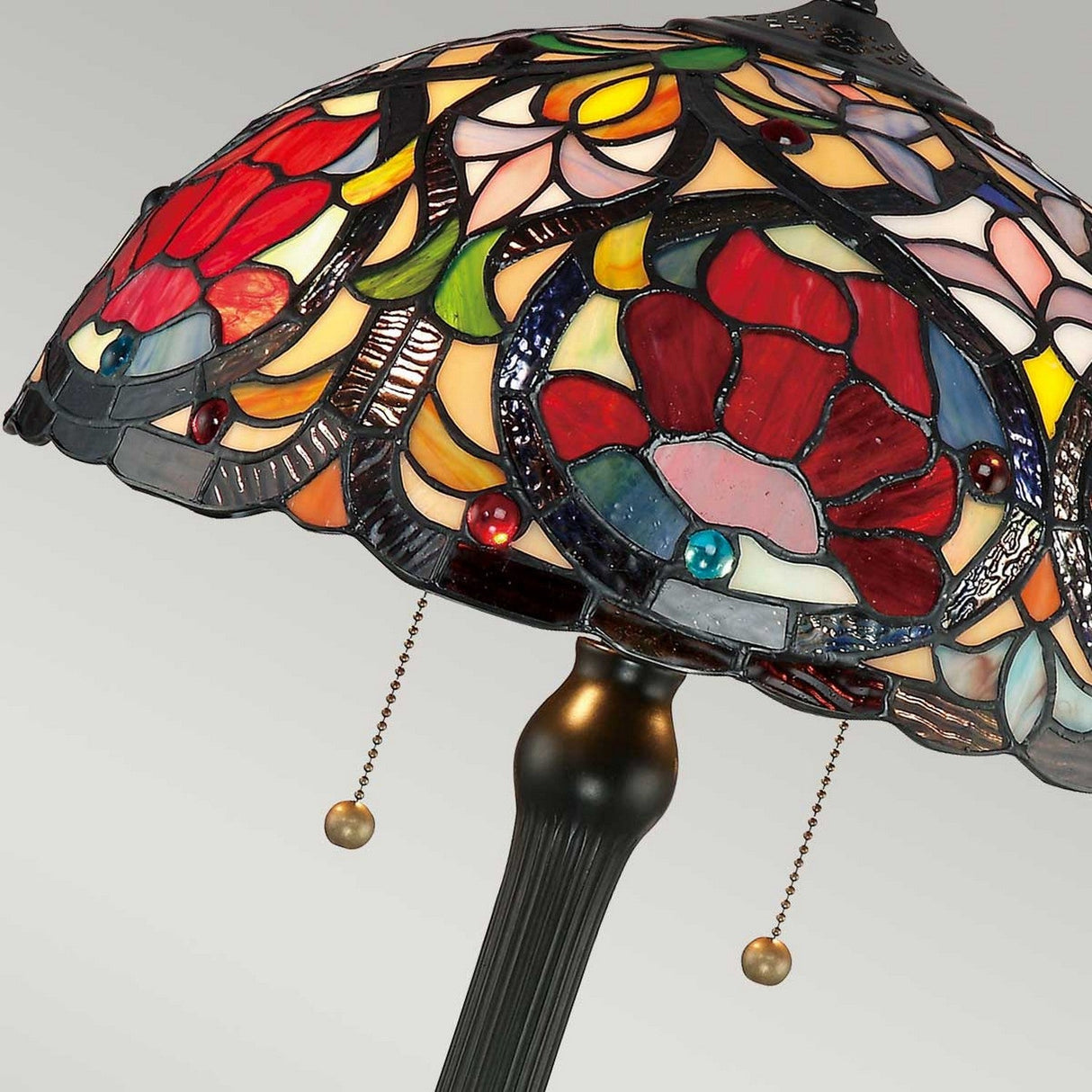 Close-up of the Larissa 2 Light Tiffany Table Lamp with an art glass shade showcasing a vibrant floral pattern in red, yellow, blue, and green. The vintage bronze base features two pull chains for convenient light adjustment.