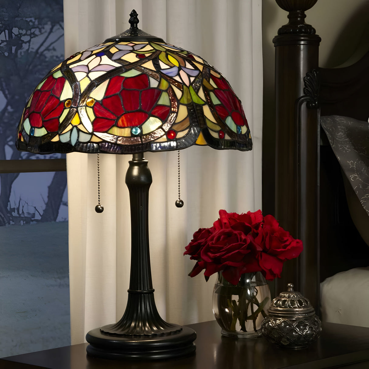 A Larissa 2 Light Tiffany Table Lamp with a floral art glass shade in red, yellow, and white rests on a bedside table. Its vintage bronze base casts a warm glow. Nearby, a small vase with red roses sits beside a metallic decorative container.