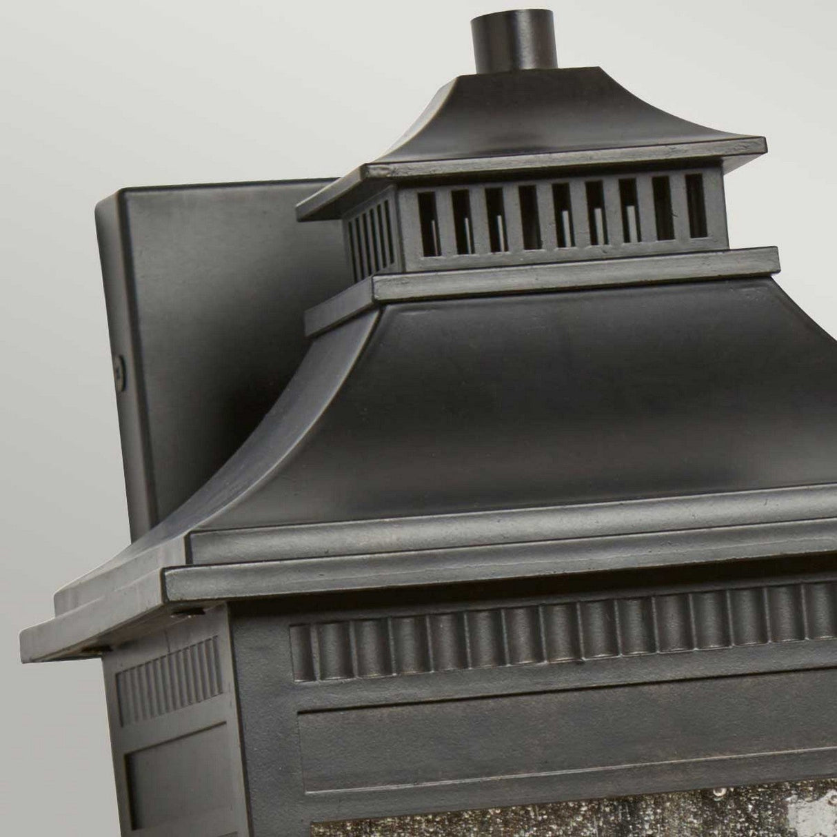 Close-up of the Livingston Outdoor 3 Light Large Wall Lantern in an elegant Imperial Bronze finish, featuring a pagoda-style roof with a detailed, tiered design and vented top. This lantern showcases seedy glass panels as it is affixed to a gray wall.