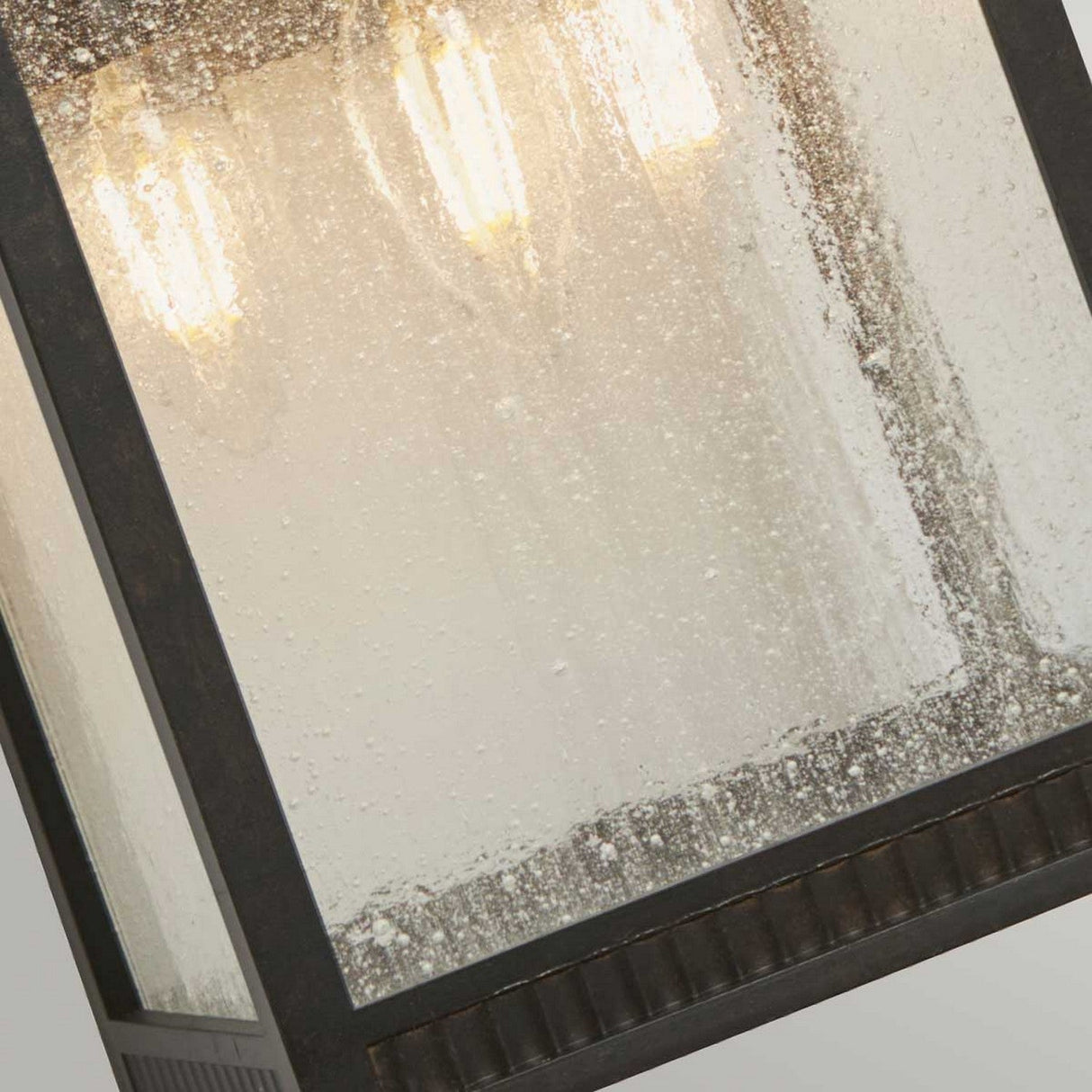 A close-up of the Livingston Outdoor 3 Light Large Wall Lantern - Imperial Bronze reveals its metal frame and textured, seedy glass. Inside, three light bulbs gently cast a warm glow through the glass. The lantern exudes a rustic and weathered charm.