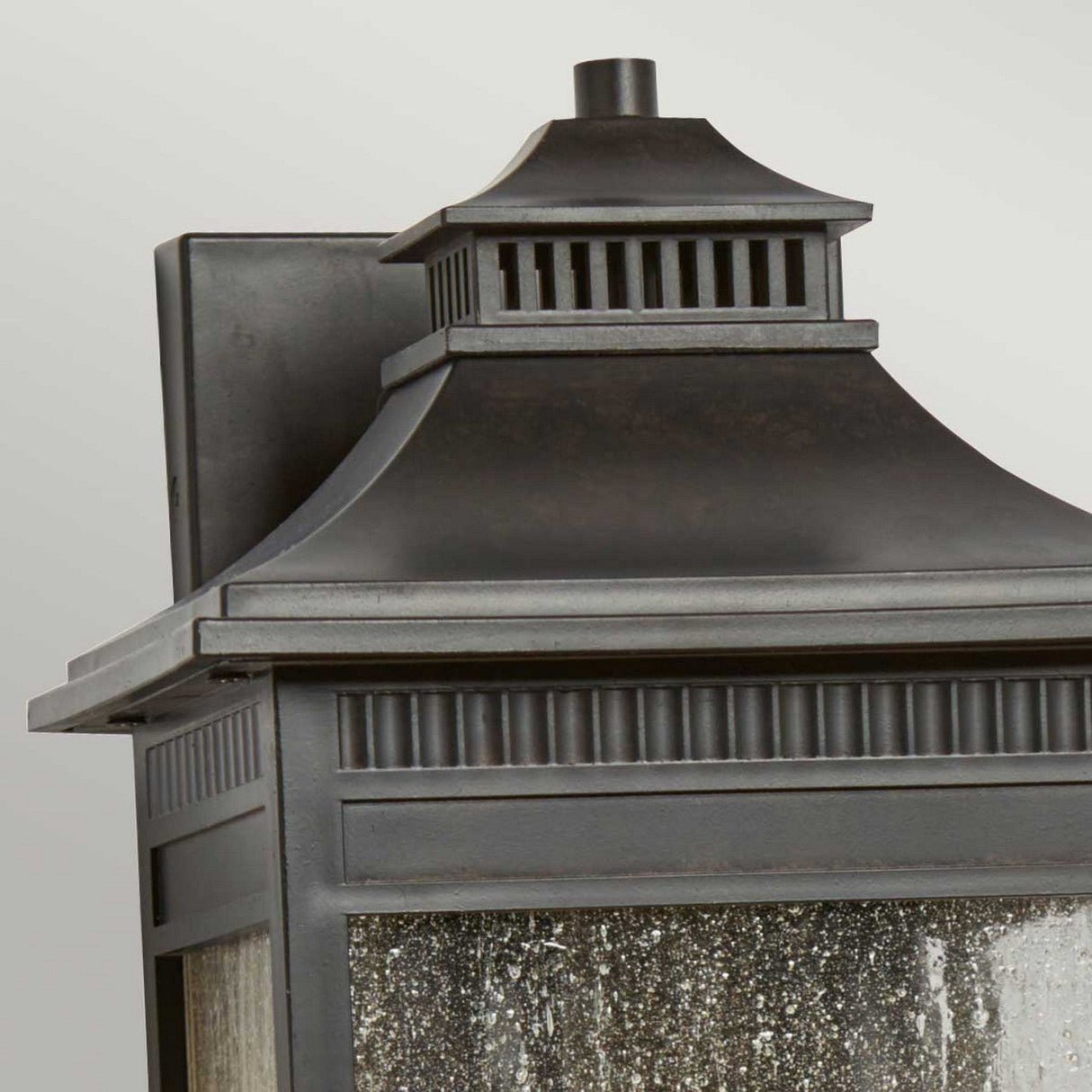 A close-up of the Livingston Outdoor 2 Light Medium Wall Lantern in Imperial Bronze showcases its dark metal finish and decorative pagoda-style roof. The lantern is adorned with vertical and horizontal grill patterns and features textured glass panes. The plain light gray background enhances its coastal design appeal.