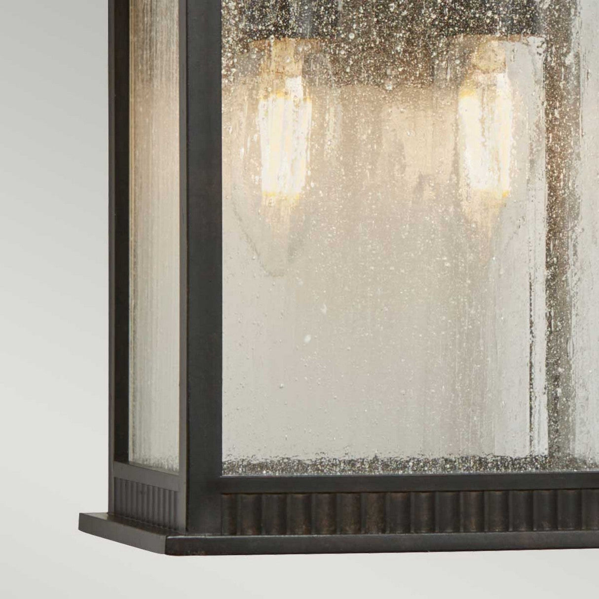 A close-up view of the Livingston Outdoor 2 Light Medium Wall Lantern showcases its black-framed design paired with textured glass panels, housing two glowing vintage-style bulbs. The lantern's imperial bronze finish adds a modern simplicity that ideally complements coastal design themes.