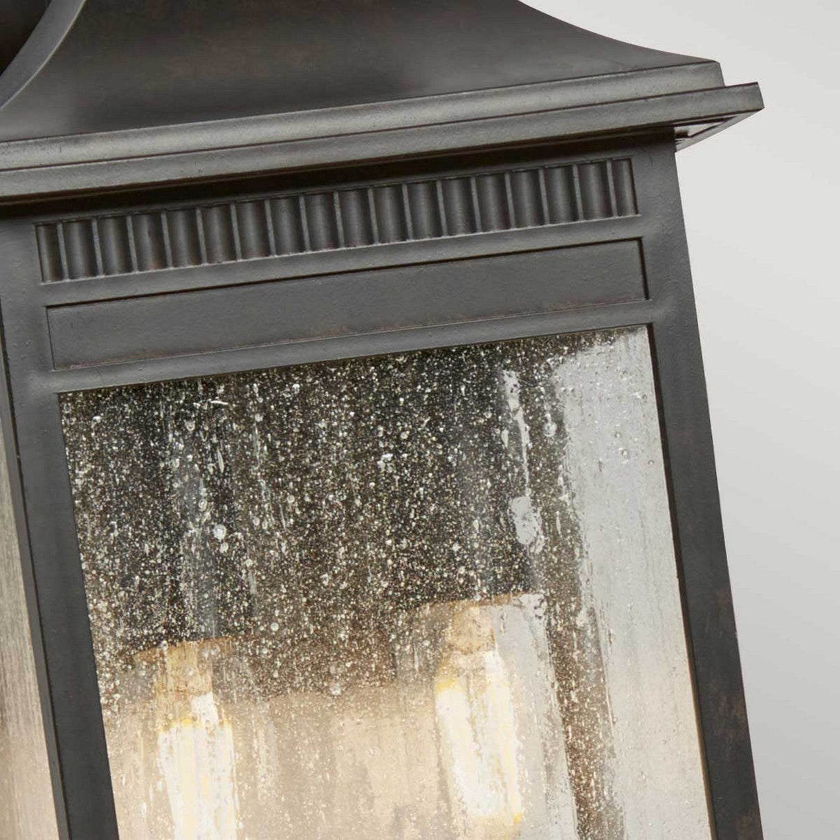 A close-up of the Livingston Outdoor 2 Light Medium Wall Lantern in an Imperial Bronze finish reveals raindrops on a textured glass panel. The light inside emits a soft glow, beautifully contrasting with the dark frame and embodying a subtle coastal design.