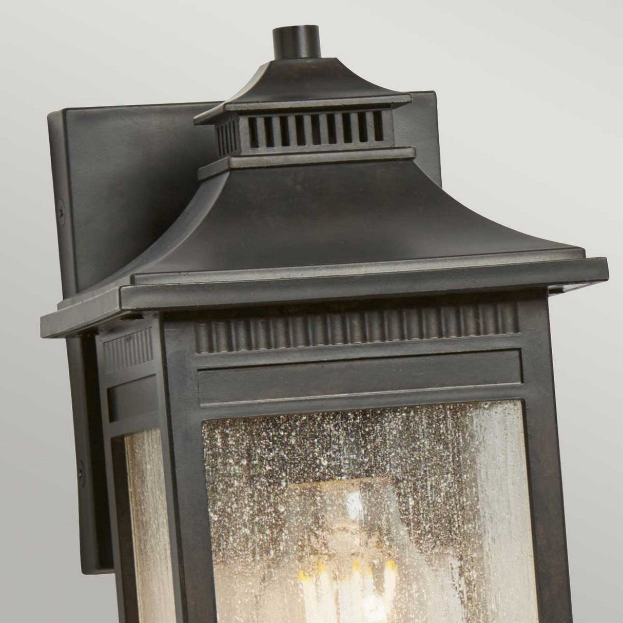 A close-up of the Livingston Outdoor 1 Light Small Wall Lantern in Imperial Bronze, featuring a traditional square design and seedy glass panels. The lamp is illuminated, casting a warm glow through the speckled glass. The background is a plain light gray.