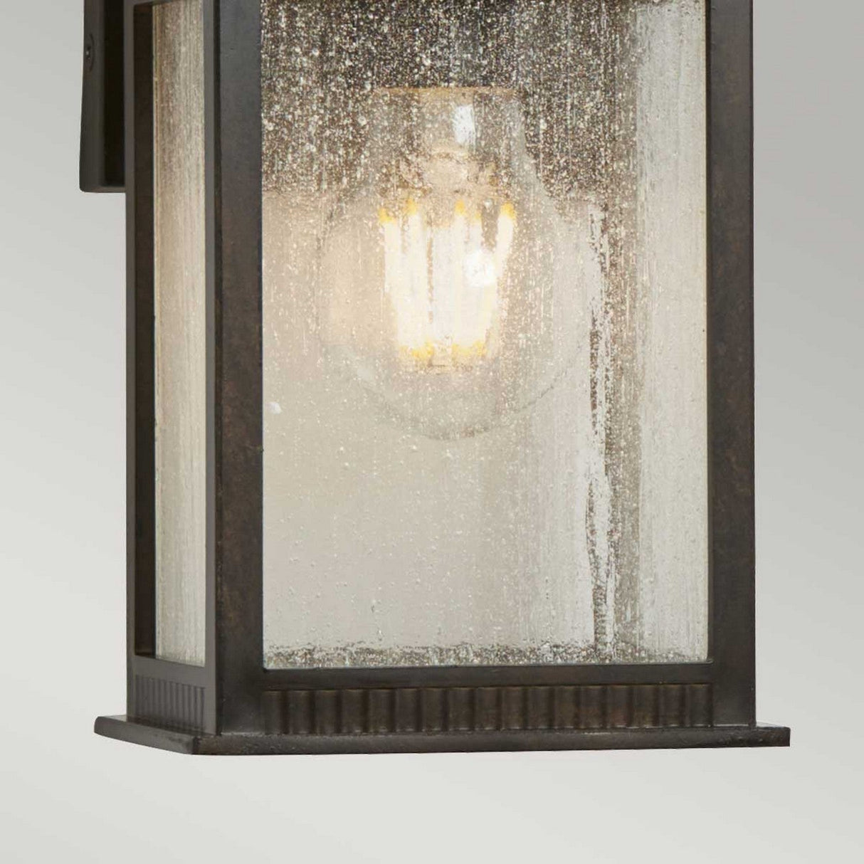 A close-up of the Livingston Outdoor 1 Light Small Wall Lantern reveals its seedy glass case and glowing light bulb that bathes the fixture in a warm glow. Its frame, finished in Imperial Bronze, adds a vintage touch.