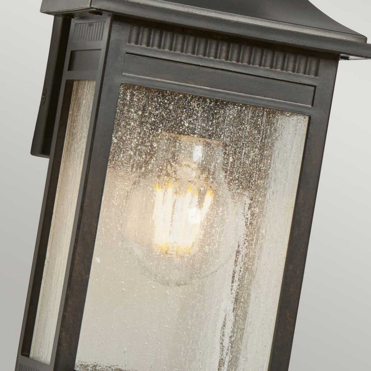 A close-up of the Livingston Outdoor 1 Light Small Wall Lantern in Imperial Bronze reveals its glowing bulb inside. The textured seedy glass, which is slightly frosted, diffuses the light beautifully. The fixture features a modern rectangular design with a matte black finish.