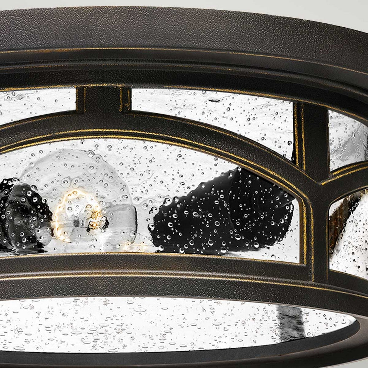 A close-up of the Marblehead Outdoor 2 Light Flush in Palladian Bronze showcases its circular design with clear glass panels, revealing droplets of water on the glass and a partially visible light bulb that creates an illuminated effect.