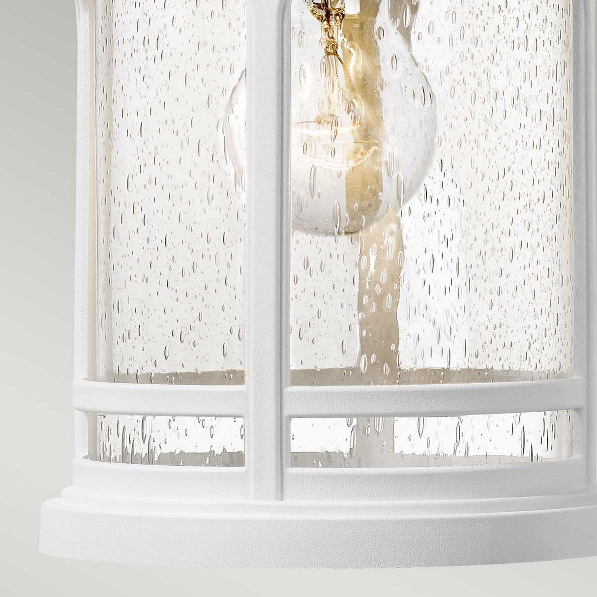Close-up of the Marblehead Outdoor 1 Light Medium Wall Lantern - White, showcasing its white frame and clear seedy glass panels. The raindrop-textured glass surrounds a glowing bulb that generates a warm ambiance, while the fixture remains weather-resistant for enduring outdoor appeal.