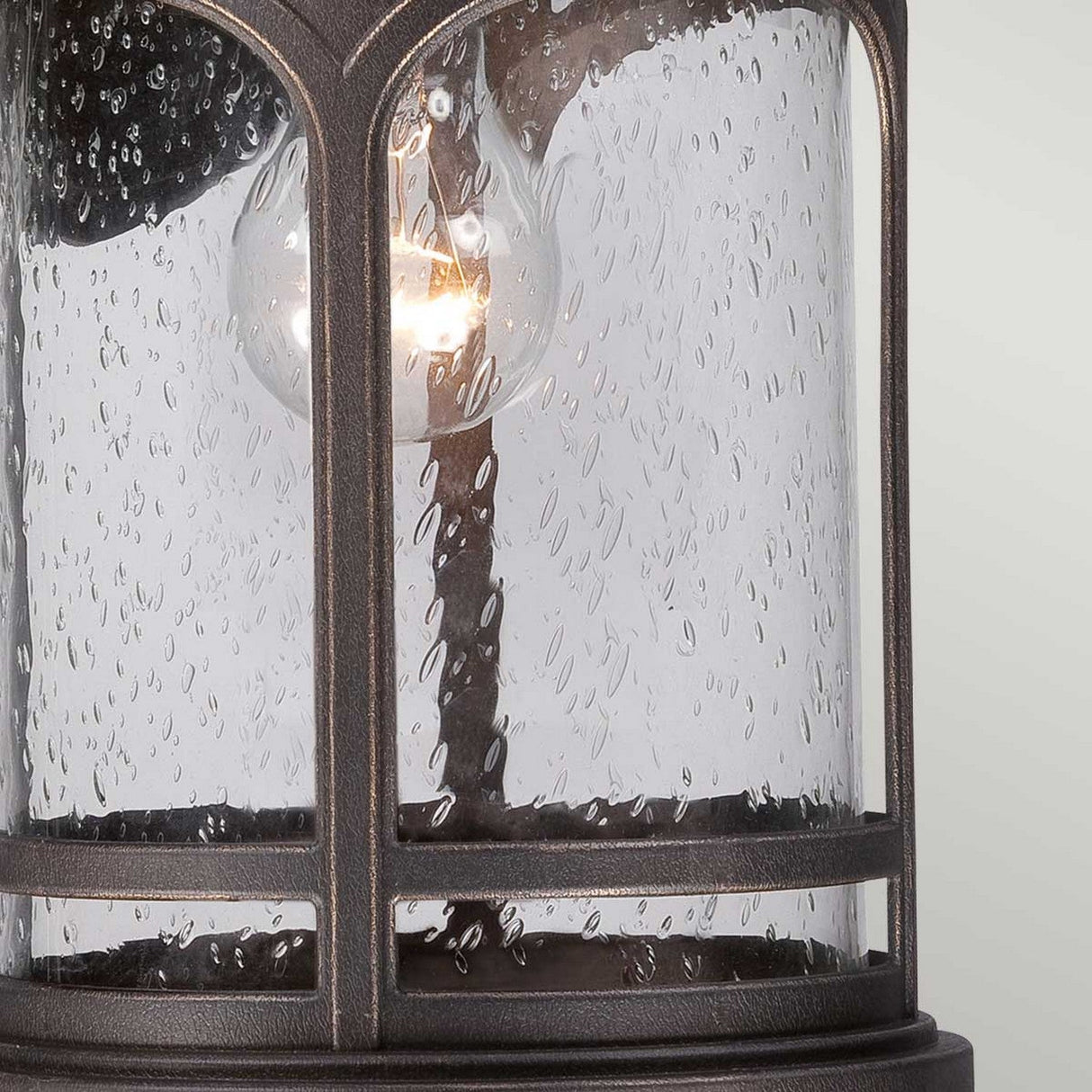 A close-up of the Marblehead Outdoor 1 Light Medium Wall Lantern in Palladian Bronze reveals its vintage style and rustic finish. Its weather-resistant glass has a textured, bubbled surface, and a glowing light bulb inside creates a warm ambiance.