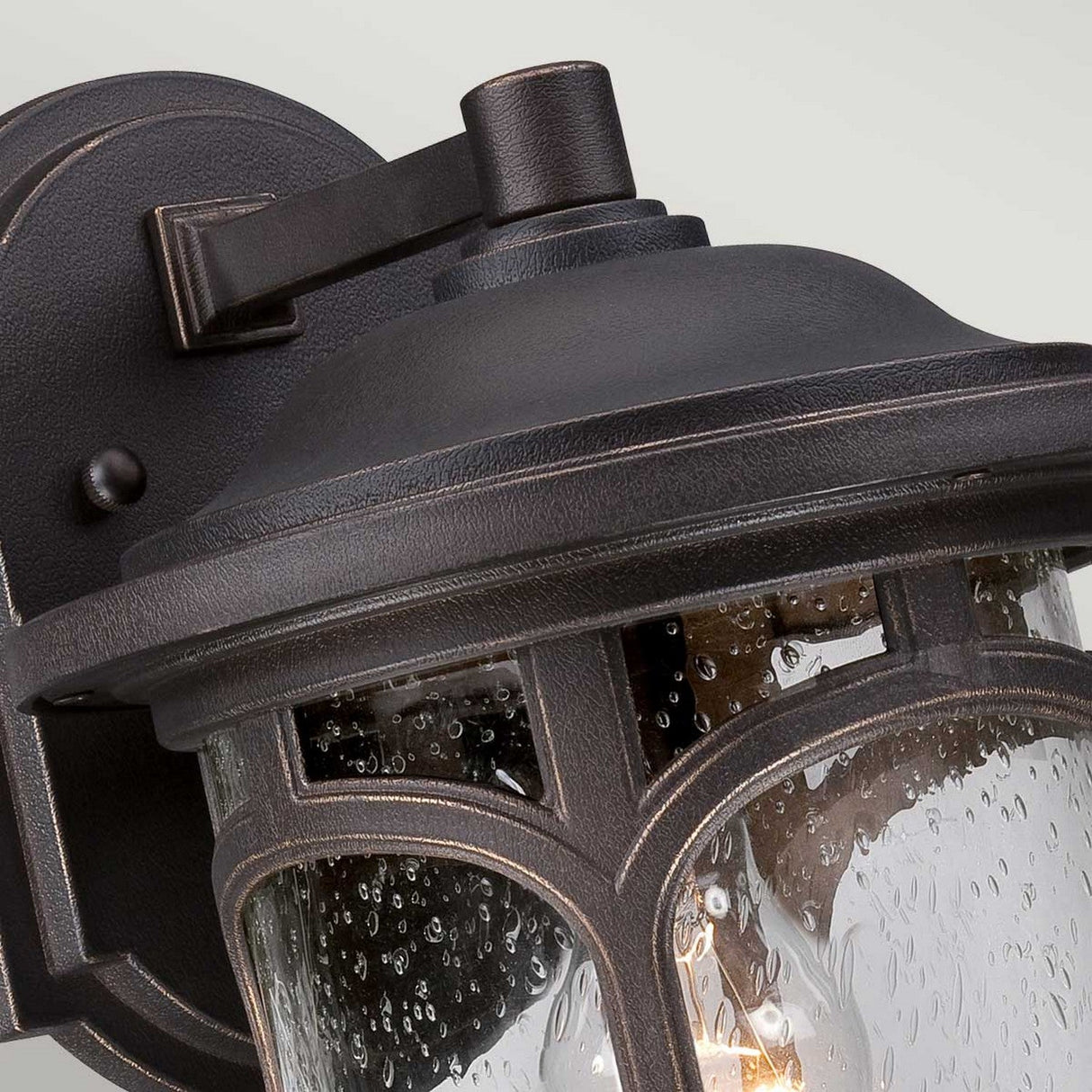 Close-up of the Marblehead Outdoor 1 Light Medium Wall Lantern in Palladian Bronze. This vintage-style lamp showcases a black metal frame with a clear glass enclosure adorned with water droplet patterns, suggesting recent rain. Weather-resistant, it emits a warm glow from within.