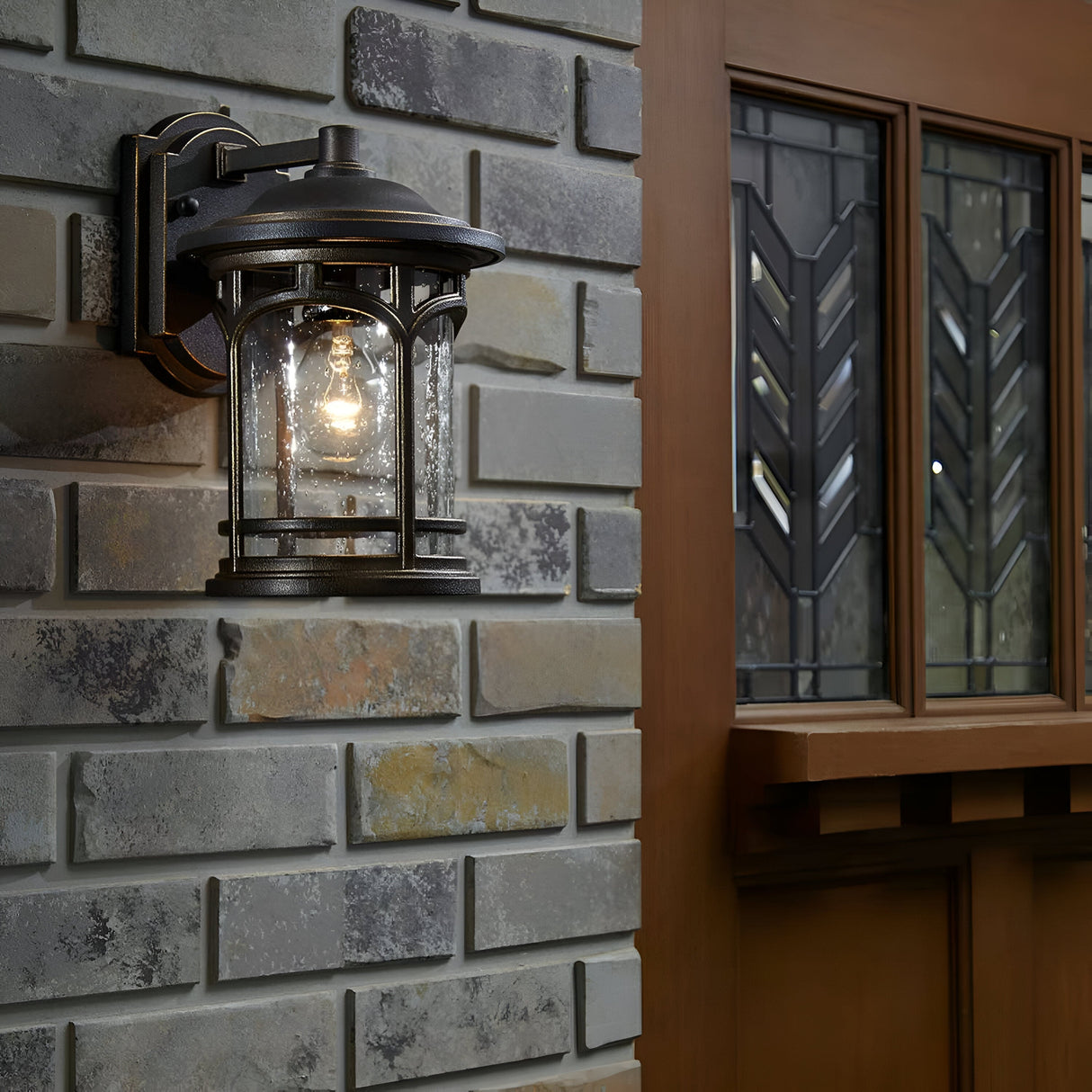 The Marblehead Outdoor 1 Light Medium Wall Lantern in Palladian Bronze, designed with a vintage style and built to withstand weather, is illuminated and mounted on a textured brick wall. In the background, a wooden door with decorative glass panels is partially visible.