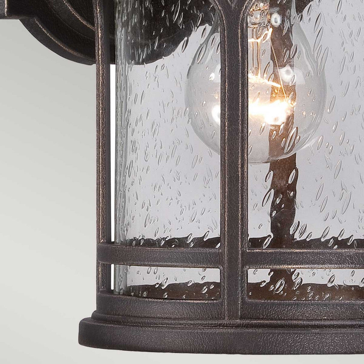A close-up of the Marblehead Outdoor 1 Light Small Wall Lantern in Palladian Bronze showcases its clear seedy glass panels, through which a glowing light bulb is visible, illuminating the textured glass. The design includes curved metal accents, adding to its classic appeal.