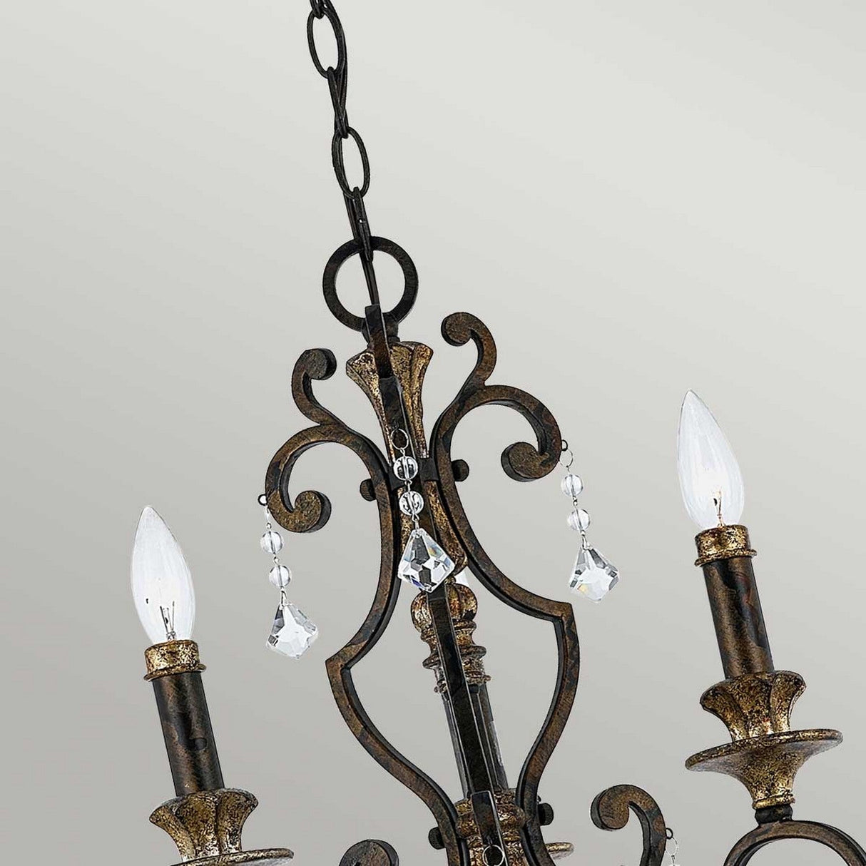 Close-up of the Marquette 3 Light Chandelier - Bronze, showcasing its ornate design with three candle-like lights topped with flame-shaped bulbs. The chandelier features intricate metalwork in a bronze finish and gleaming crystal drops hanging from its center. The background is a plain light color.