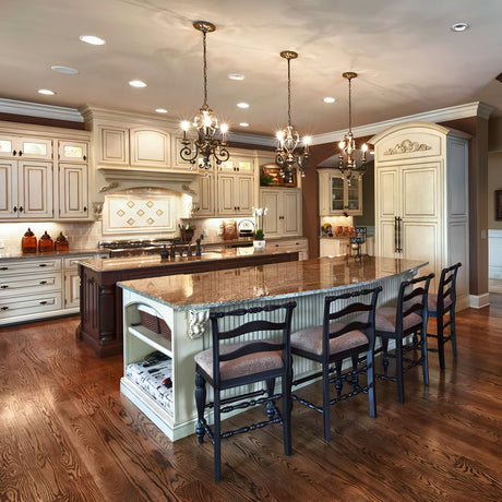 A luxurious kitchen showcases the Marquette 3 Light Chandelier in bronze, adorned with crystal drops, above a large island featuring a brown granite countertop and four chairs. The space is further enhanced by elegant ivory cabinetry, wooden floors, and tasteful decorative elements, perfectly blending elegance with functionality.