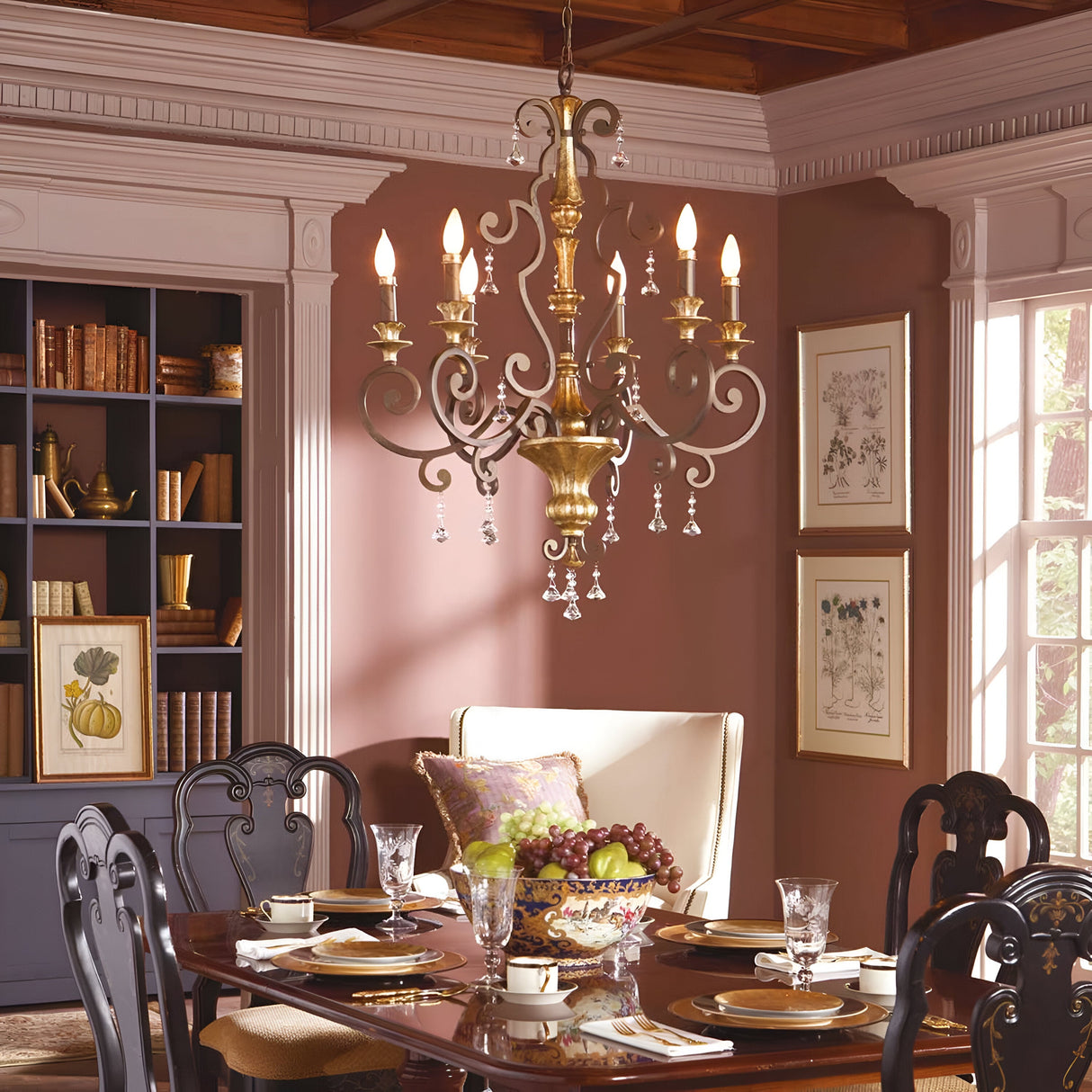 A dining room featuring the sophisticated Marquette-A 6 Light Chandelier in bronze, adorned with crystal drops. Below it is a polished wooden table set for six, complete with plates, glassware, and a fruit bowl. The room also showcases dark wood chairs, a bookshelf, framed artwork, and a large window that lets in natural light.