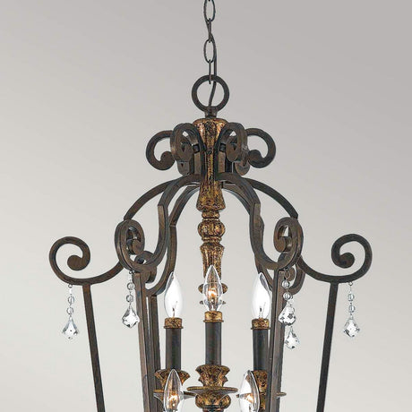 Introducing the Marquette-B 6 Light Chandelier - Bronze: a decorative masterpiece with an ornate metal frame in rich bronze, featuring scrolling designs and crystal drops. The fixture holds multiple candle-shaped lights, crafting an elegant and vintage appearance.