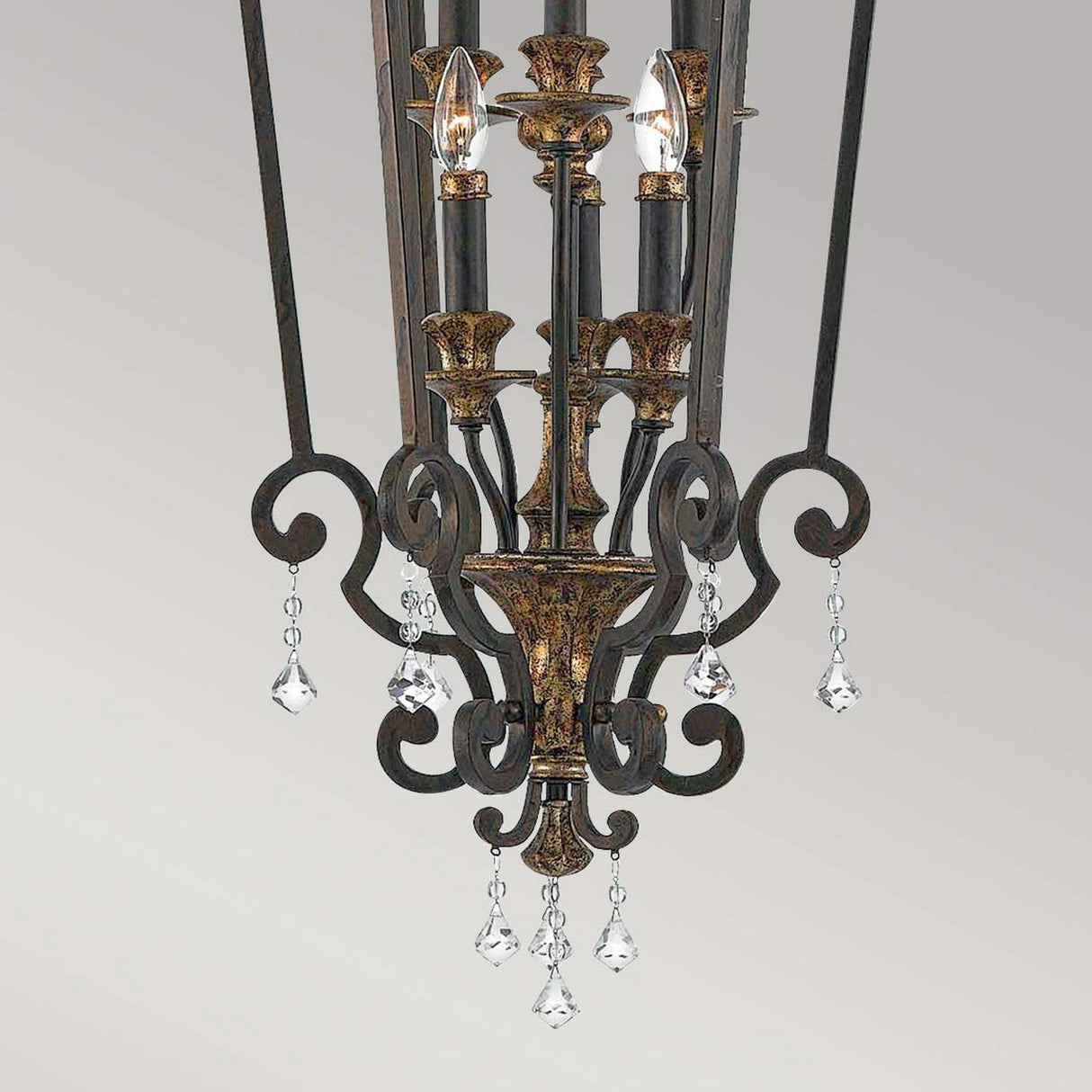 The Marquette-B 6 Light Chandelier - Bronze is an elegant fixture showcased by its black and gold metal finish, intricate scrollwork, and hanging crystal accents. This bronze chandelier features six candle-style lights adorned with sparkling crystal drops.
