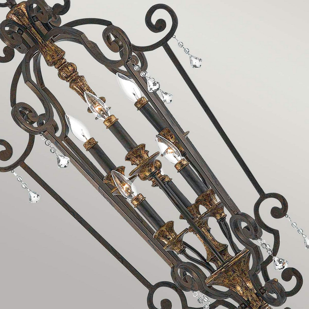 The Marquette-B 6 Light Chandelier in Bronze showcases an intricate wrought iron design with elegant scrollwork and decorative elements. It features multiple candle-shaped lights and delicate crystal drops, all set against a neutral background.