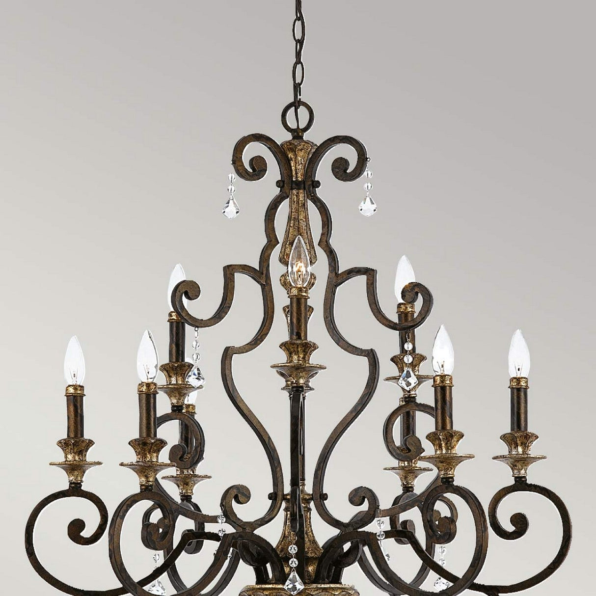 A Marquette 9 Light Chandelier in bronze, featuring elegant crystal drops hanging on a chain against a plain background. The design boasts intricate scrollwork and nine candle-style lights with an antique bronze finish.