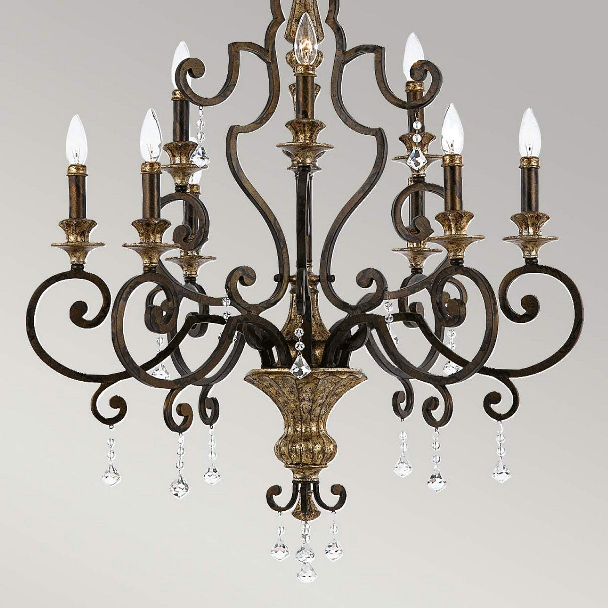 The Marquette 9 Light Chandelier in bronze combines vintage style with ornate dark metal scrollwork and features nine candle-style lights adorned with elegant crystal drops hanging from its arms. When illuminated, it casts a warm and inviting glow against a neutral background.