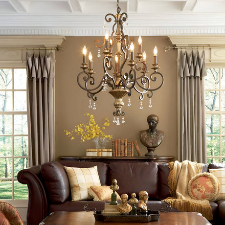 A living room with a brown leather sofa, decorative pillows, and a coffee table adorned with small busts. The Marquette 9 Light Chandelier in bronze with crystal drops hangs above. The walls feature books, a bust sculpture, and decorative vases. Beige curtains frame the tall windows elegantly.