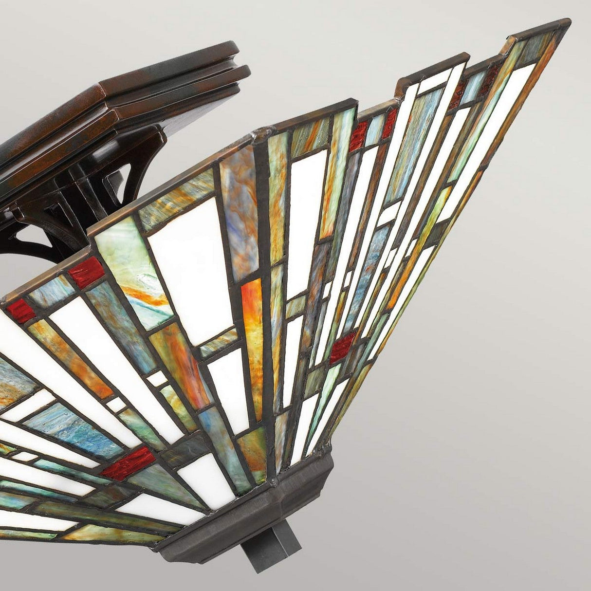 A close-up view of the Maybeck 2 Light Tiffany Flush - Valiant Bronze lamp shade reveals its angular fan-like design, highlighting the exquisite handcrafted artistry. The geometric pattern in shades of white, green, red, and amber captures a visually striking effect that is reminiscent of a traditional Tiffany-style ceiling light.