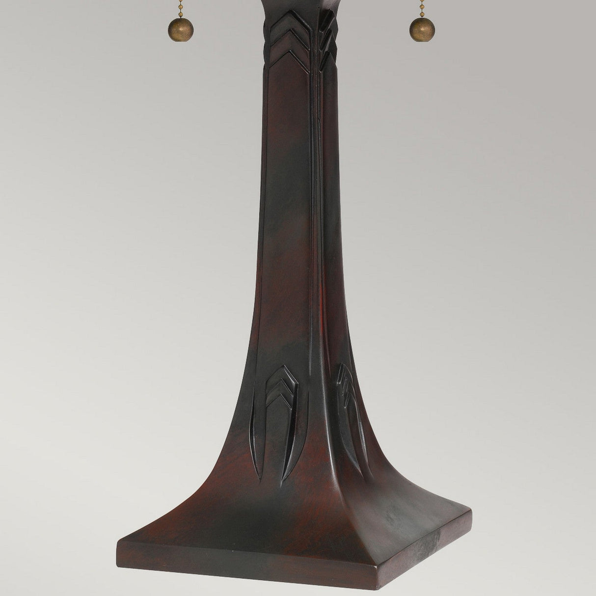 The Maybeck 2 Light Tiffany Table Lamp in Valiant Bronze features a dark wooden lamp base with a square, wide bottom and vertical carved decorative lines that extend upward. Two pull chains with small round weights hang from the top against a plain light gray background.