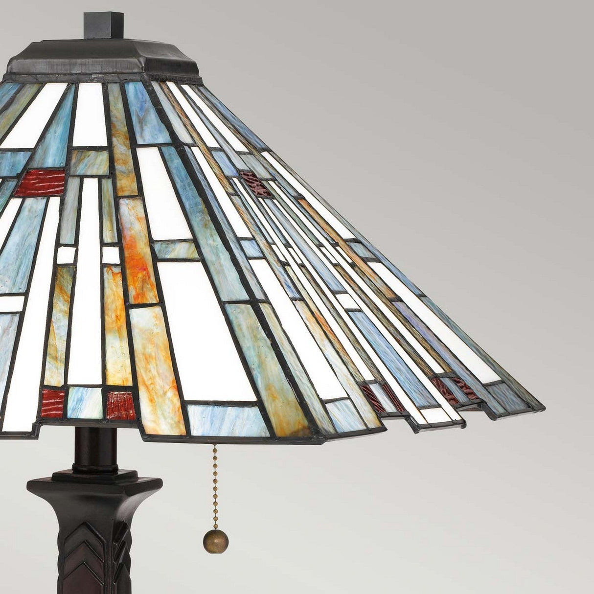 The Maybeck 2 Light Tiffany Table Lamp - Valiant Bronze showcases a stained glass lampshade with geometric patterns in white, blue, and amber hues. It features a dark base with a Valiant Bronze finish and is equipped with a dangling chain pull switch, allowing its handcrafted glass design to stand out beautifully against a simple gray background.