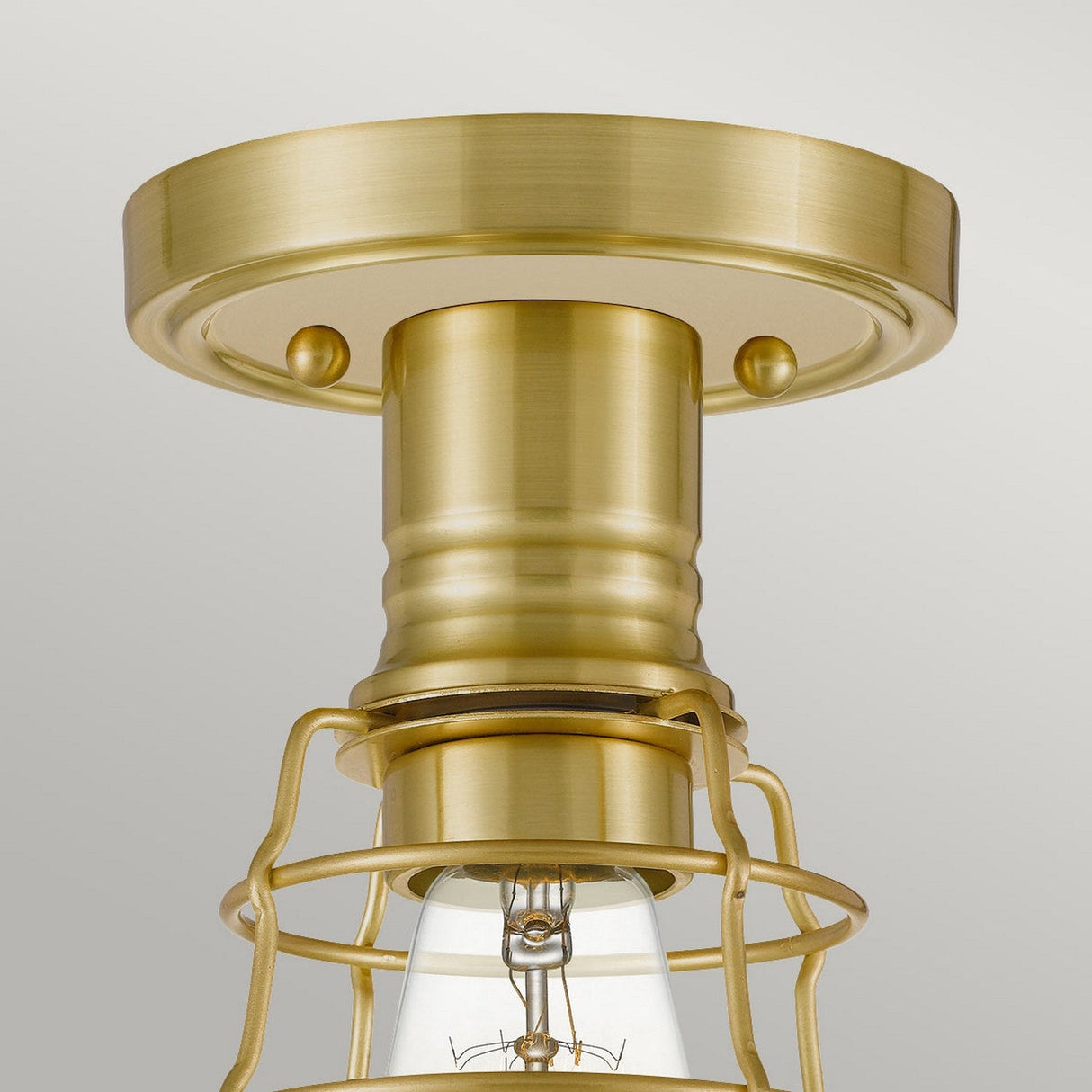 A close-up of the Mixon 1 Light Semi-Flush in Brushed Brass showcases its cylindrical base and partially visible bulb, enclosed within a protective wire cage, exemplifying an industrial light fixture style against a neutral background.