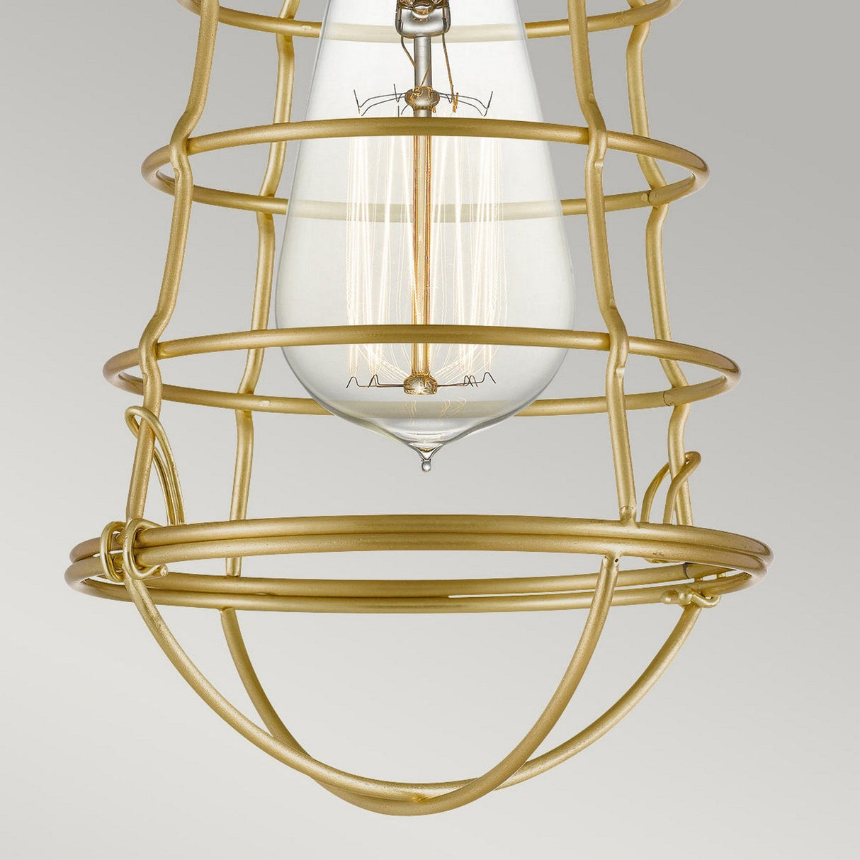 A close-up of the Mixon 1 Light Semi-Flush - Brushed Brass showcases a decorative cage design encircling a vintage-style bulb. The cage forms an elegant geometric pattern against a neutral background, epitomizing modern industrial aesthetics.