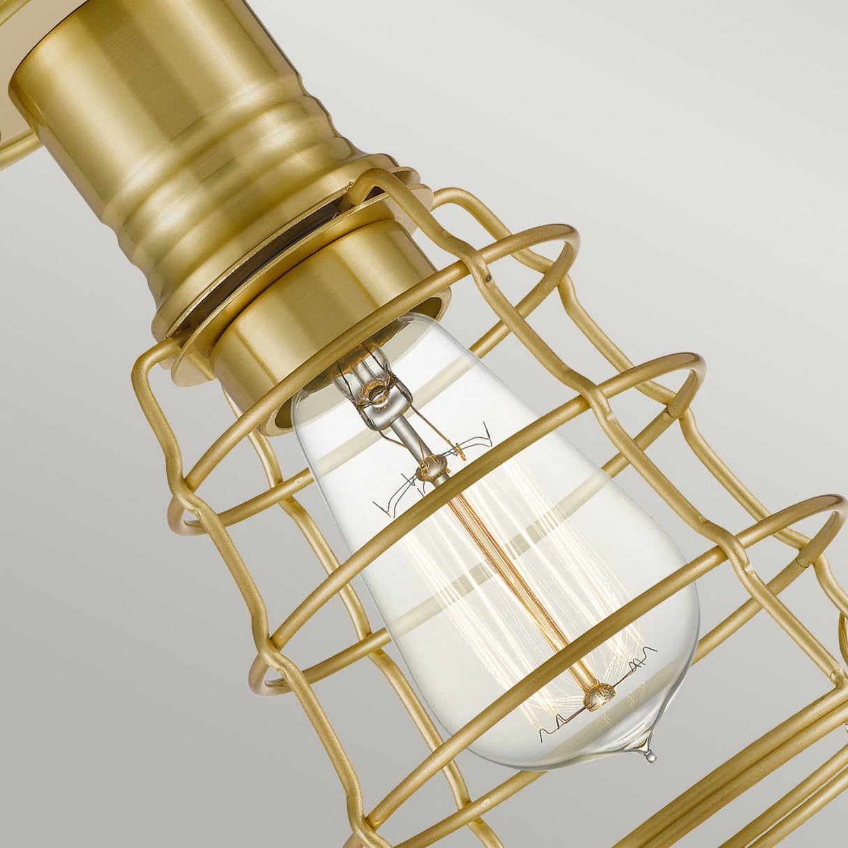 Close-up of the Mixon 1 Light Semi-Flush fixture in brushed brass, highlighting a vintage light bulb encased in a brass cage-style design. The elongated, clear bulb is surrounded by a brushed brass grid, showcasing the fixture's warm, industrial aesthetic against a plain background.