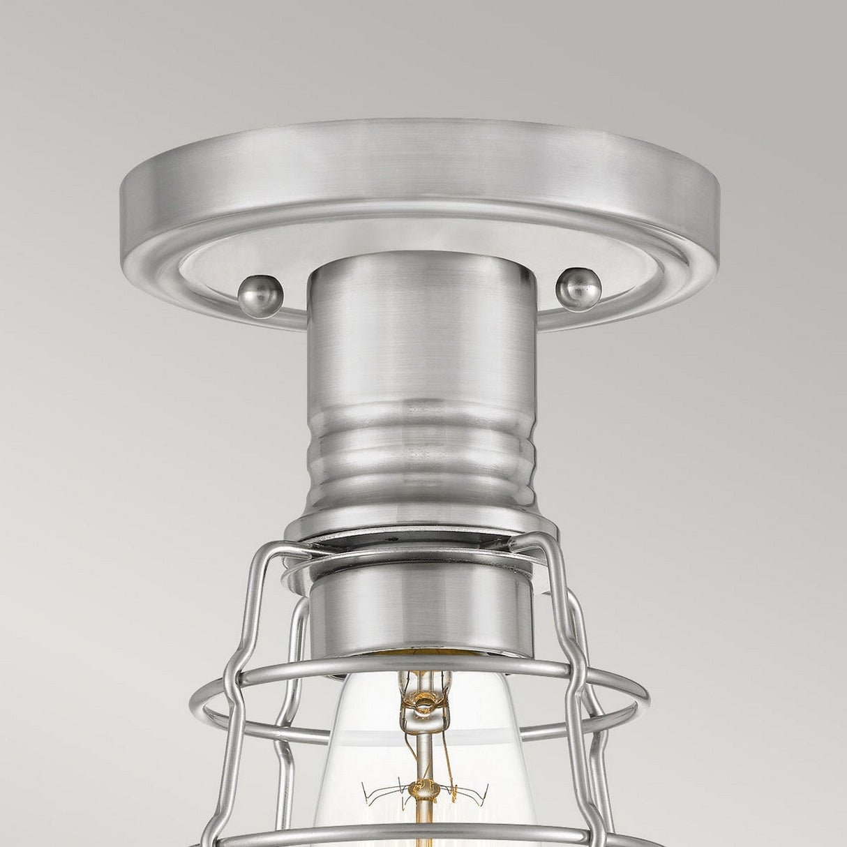 A close-up of the Mixon 1 Light Semi-Flush in brushed nickel showcases its industrial style. It highlights a cylindrical base with a wire cage surrounding a clear bulb. The plain, light gray background beautifully complements its semi-flush mount design.