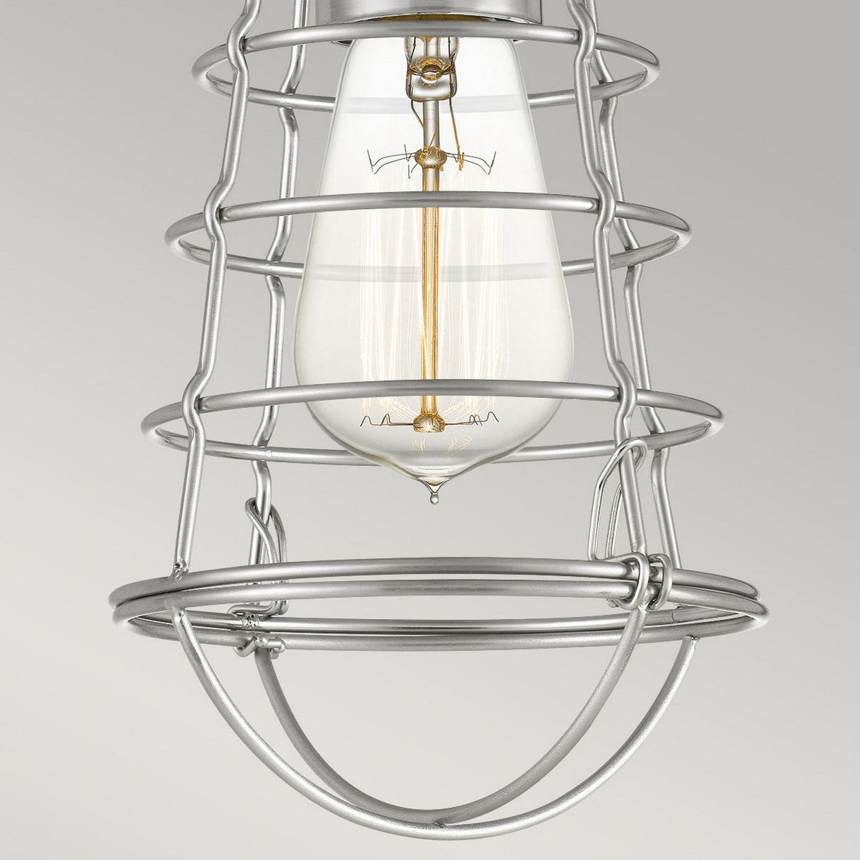A close-up of the Mixon 1 Light Semi-Flush in Brushed Nickel showcases a vintage-style bulb encased in an industrial metal cage. The brushed nickel finish with its grid-like design perfectly complements the clear glass bulb that unveils the filament inside, all set against a plain, neutral gray background.