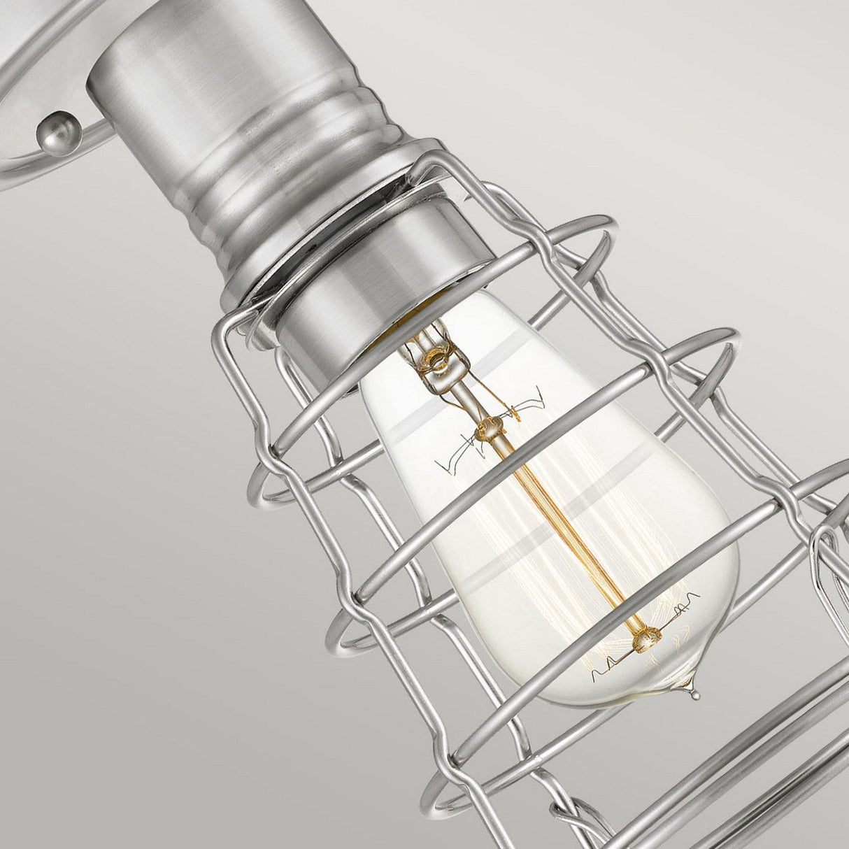 A close-up of the Mixon 1 Light Semi-Flush - Brushed Nickel reveals an industrial-style light bulb encased in a metal cage fixture. The glowing filament shines brightly against a neutral gray background, enhanced by brushed nickel accents that add a touch of elegance to its vintage charm.