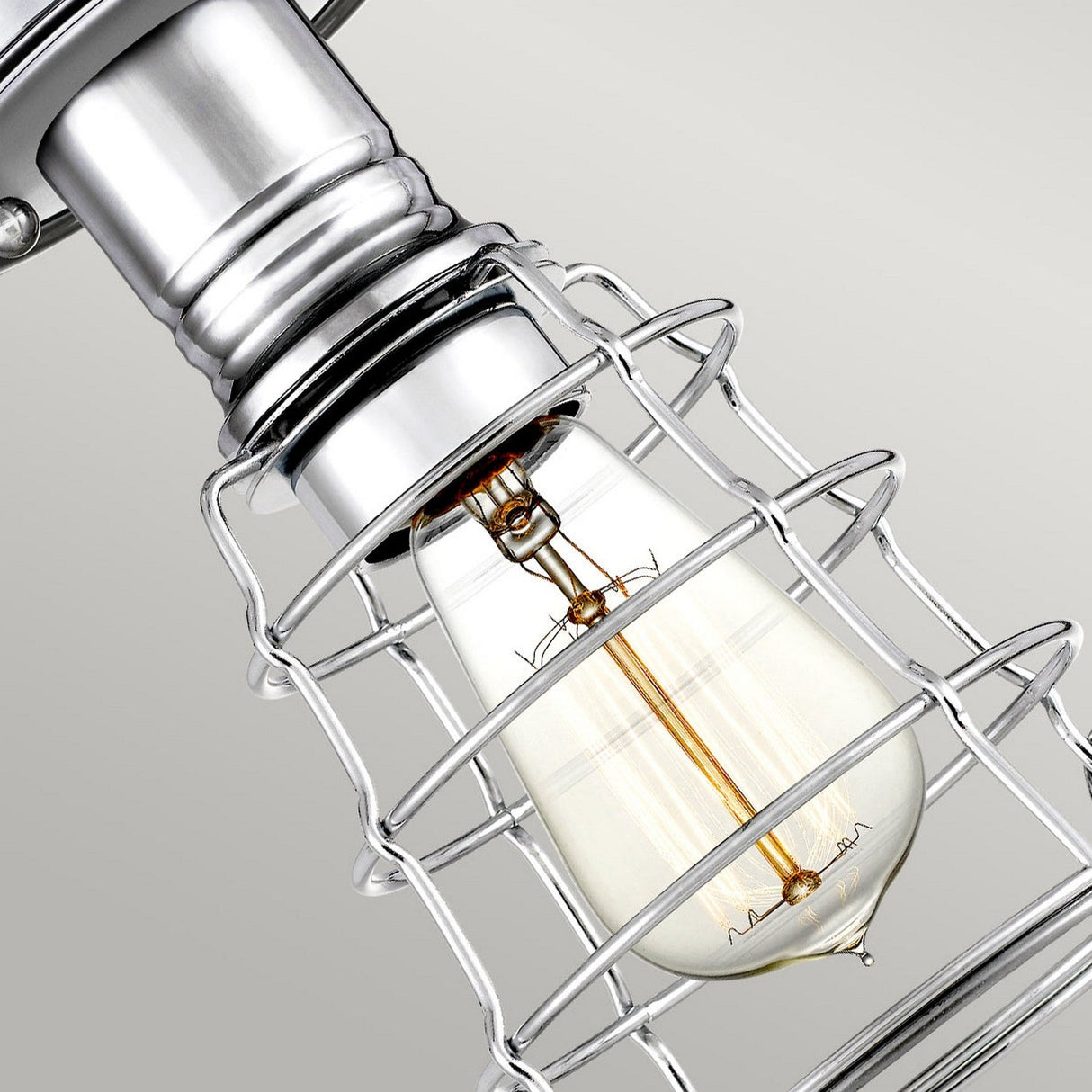 A close-up of the Mixon 1 Light Semi-Flush in polished chrome showcases a vintage-style lightbulb encased in a silver metal cage, emitting a warm glow. This fixture enhances the industrial-chic design against a plain background.