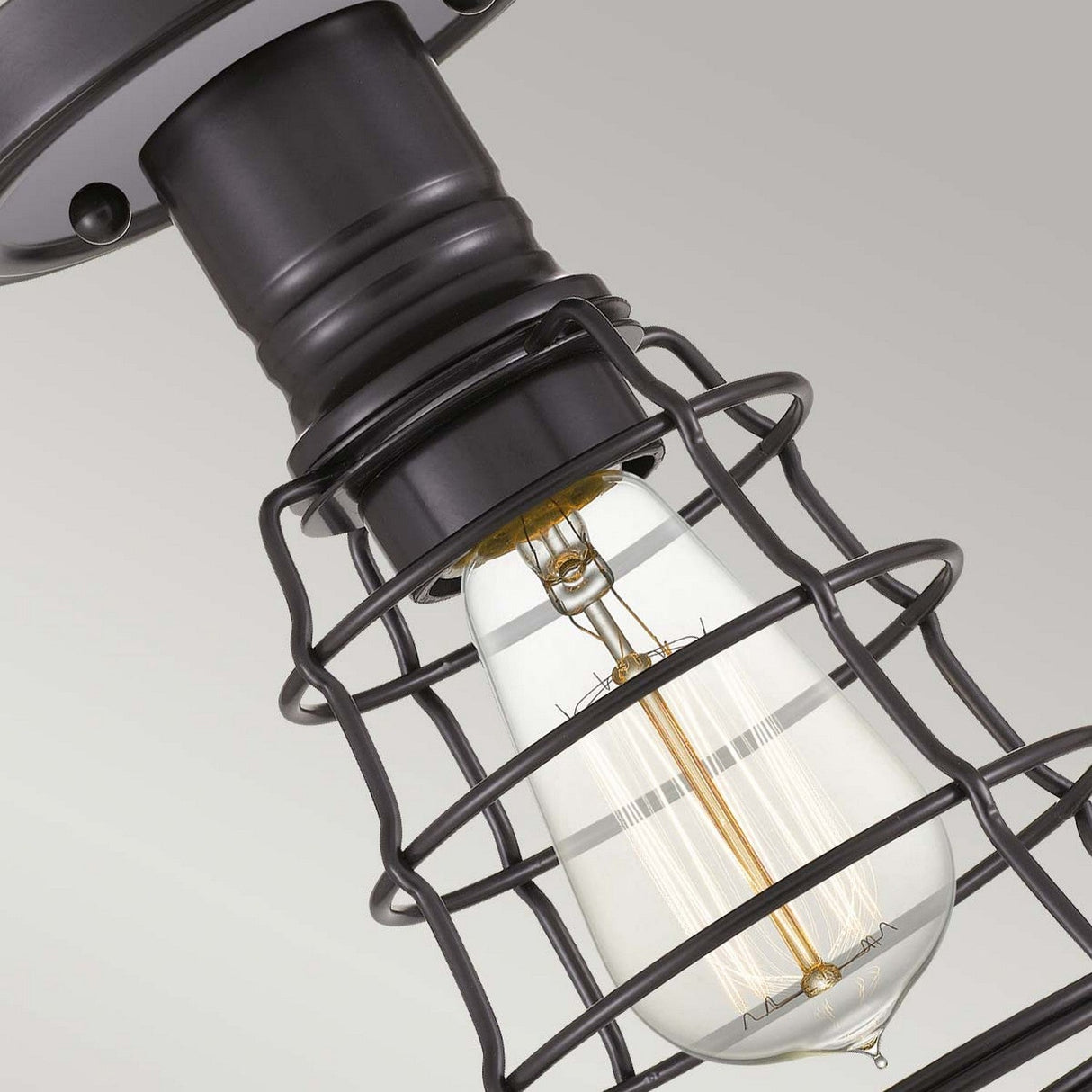 The Mixon 1 Light Semi-Flush - Palladian Bronze features a vintage Edison bulb enclosed in a black metal cage, mounted on a brushed metal base with hints of Palladian Bronze, exuding industrial appeal against a plain background.