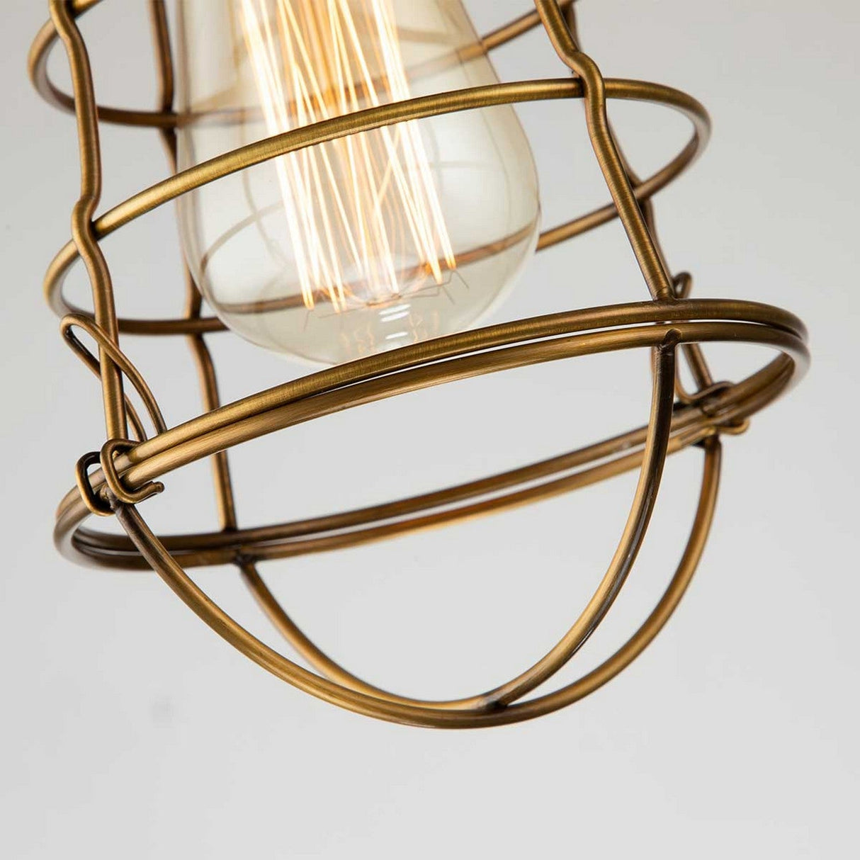 A Mixon 1 Light Semi-Flush fixture in weathered brass showcases an industrial-style bulb within a vintage-inspired bronze wire cage. The geometric pattern design emits a warm glow from the filament, set against a neutral light gray background.