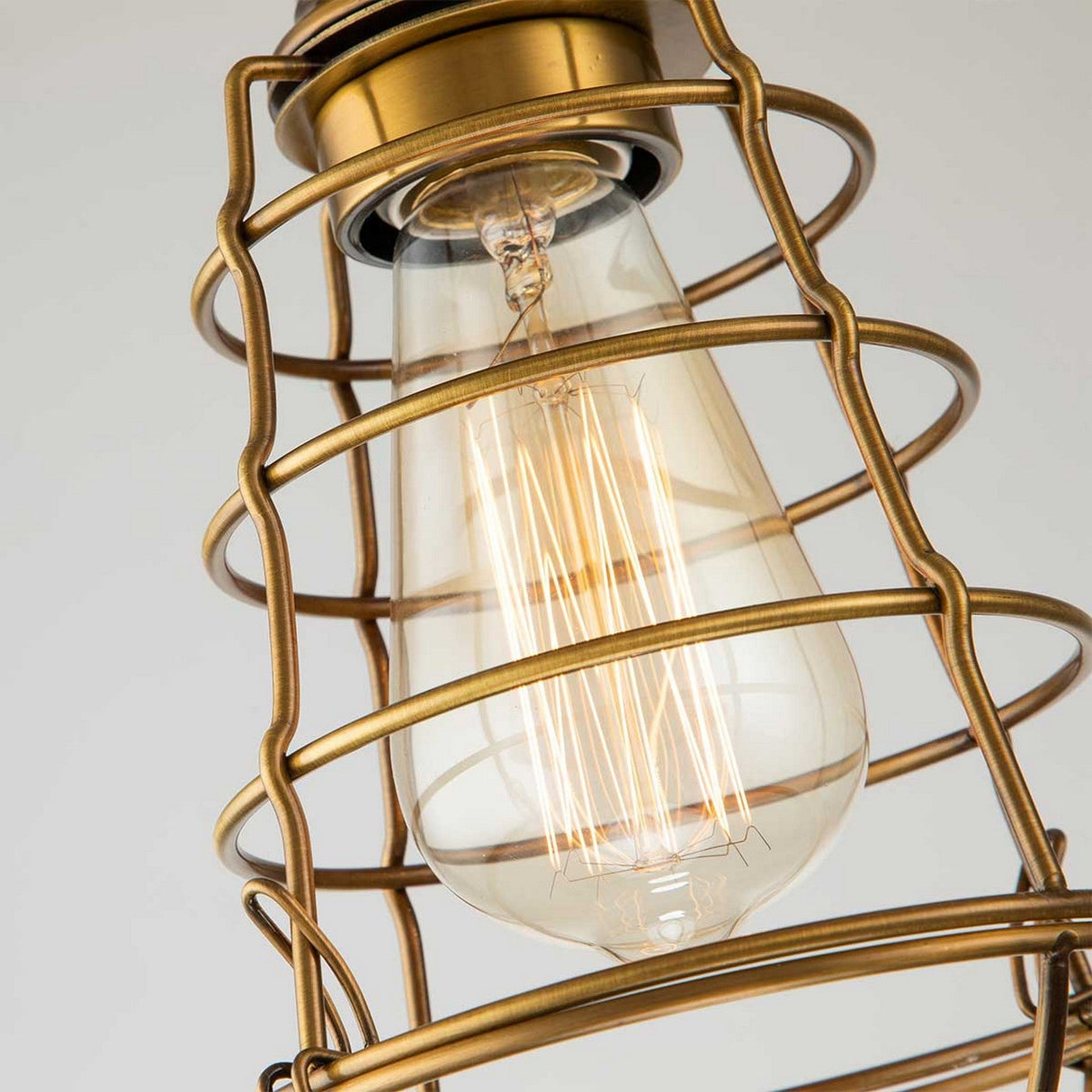 Close-up of the Mixon 1 Light Semi-Flush fixture showcasing its vintage-inspired design with a filament visible inside a brass wire cage. The industrial aesthetic is enhanced by the intricate geometric pattern of its surrounding structure, featuring a warm, weathered brass finish.