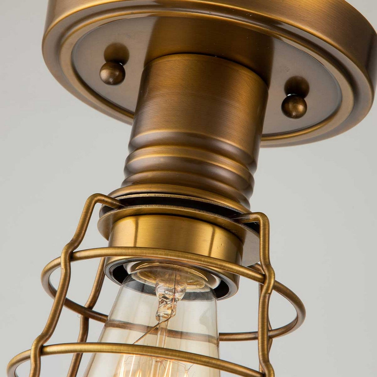 A close-up of the Mixon 1 Light Semi-Flush in weathered brass showcases its exposed bulb within a wire cage. This piece epitomizes a vintage industrial style, complete with a metallic finish and decorative screws that capture the essence of vintage-inspired lighting.