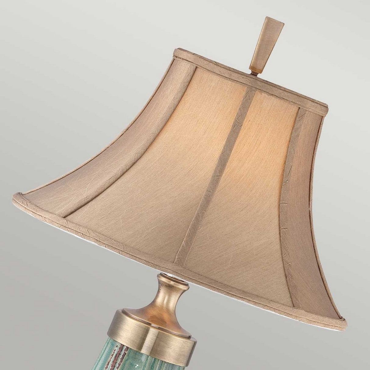 A close-up of the Monteverde 1 Light Table Lamp - Green/Aged Brass, highlighting its decorative design with a beige textured lampshade and an aged brass finish finial. The brass-colored base beautifully complements its glass body. The lampshade tilts slightly to the right against a neutral background.