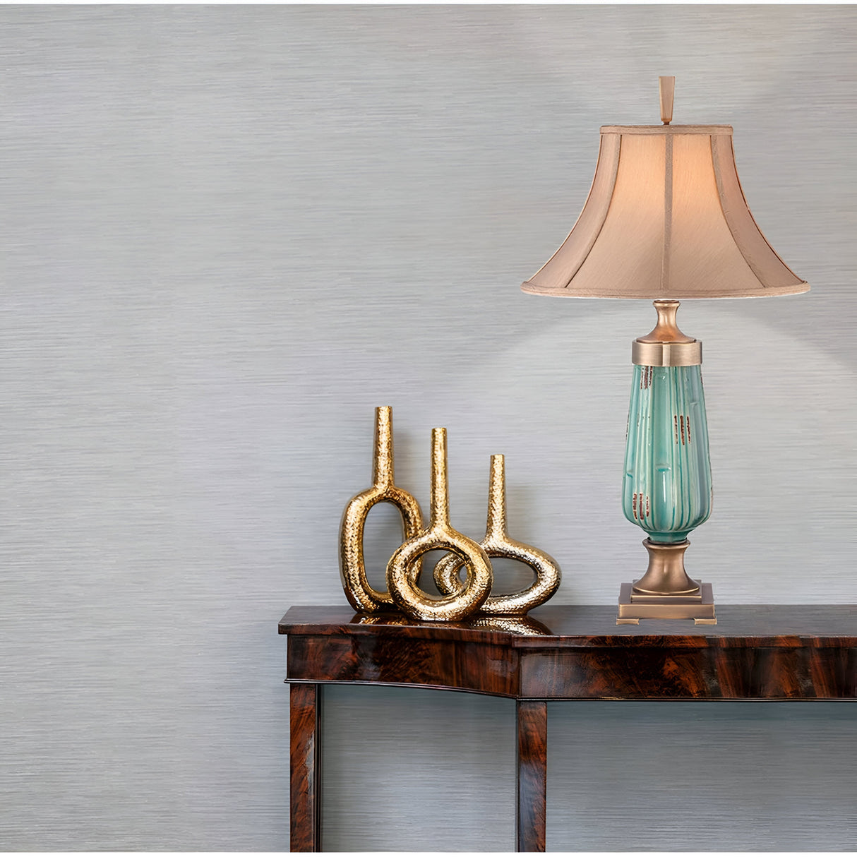 A decorative arrangement on a wooden table against a neutral wall features the Monteverde 1 Light Table Lamp with an aged brass finish and beige shade, complemented by three gold looped sculptures. The table has a glossy, dark wood finish that enhances the elegant display.