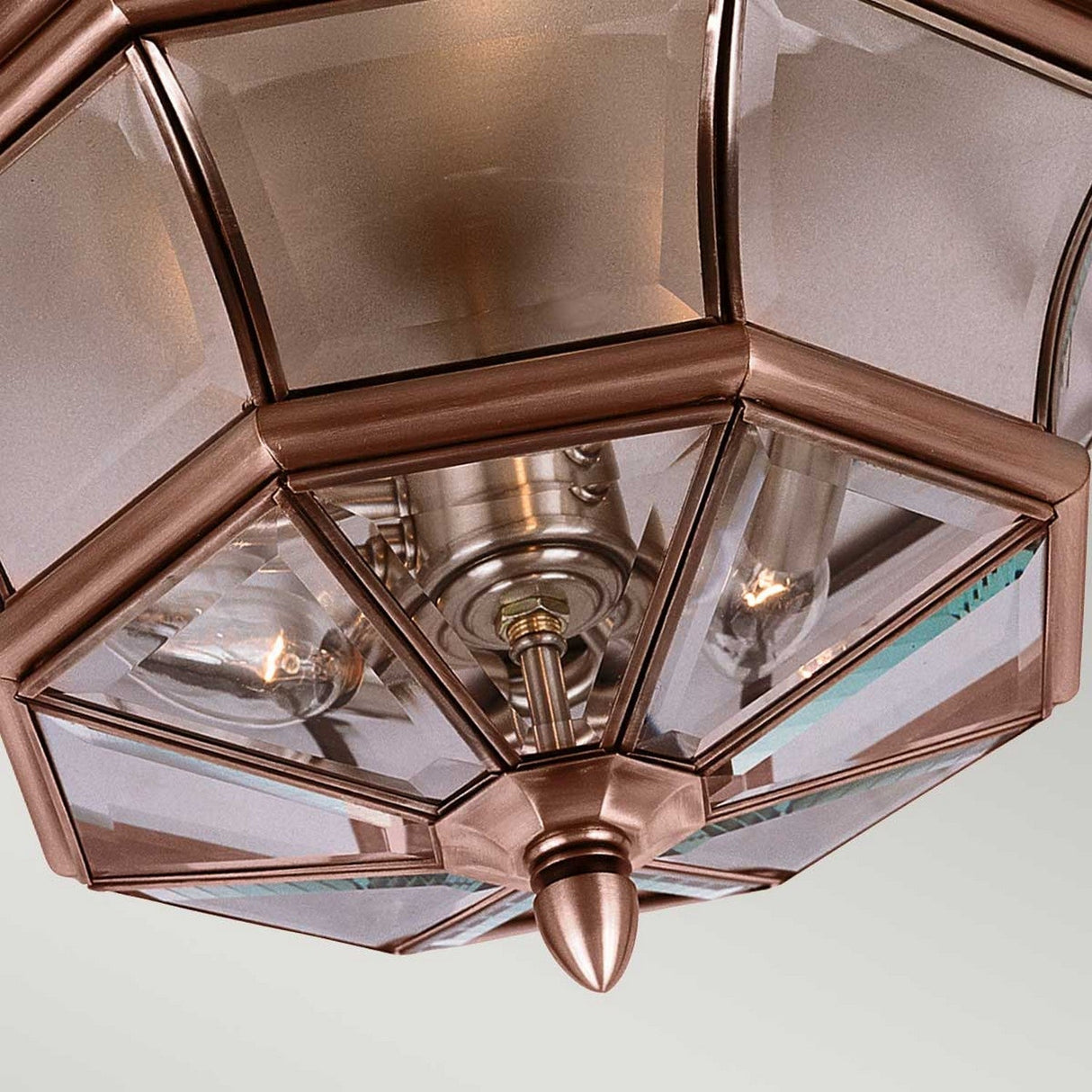 A close-up of the Newbury Outdoor 3 Light Flush - Lacquered Aged Copper reveals its geometric design, featuring beveled glass panels that showcase visible light bulbs inside. This flush mount outdoor light fixture boasts an aged copper finish and a hexagonal shape with a pointed finial at the bottom.