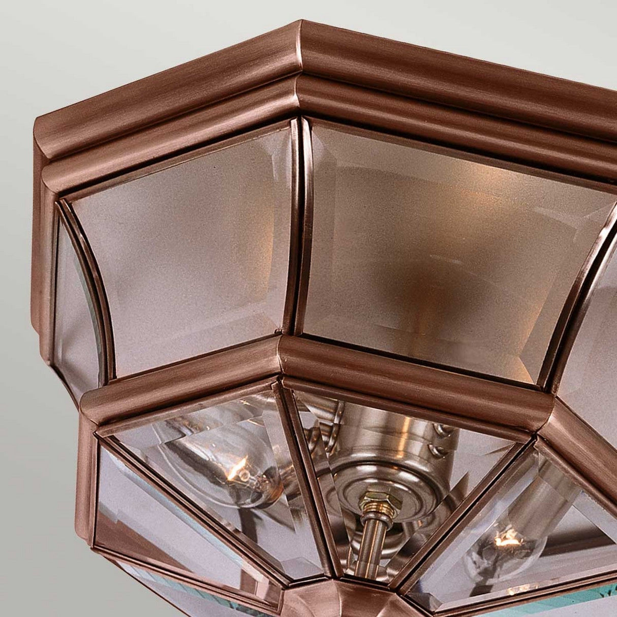 Close-up of the Newbury Outdoor 3 Light Flush ceiling light fixture with a hexagonal design featuring glass panels and an exposed bulb inside. The lacquered aged copper finish contrasts with the clear glass, resulting in a modern and elegant look that is ideal for both indoor flush mount lighting and outdoor settings.
