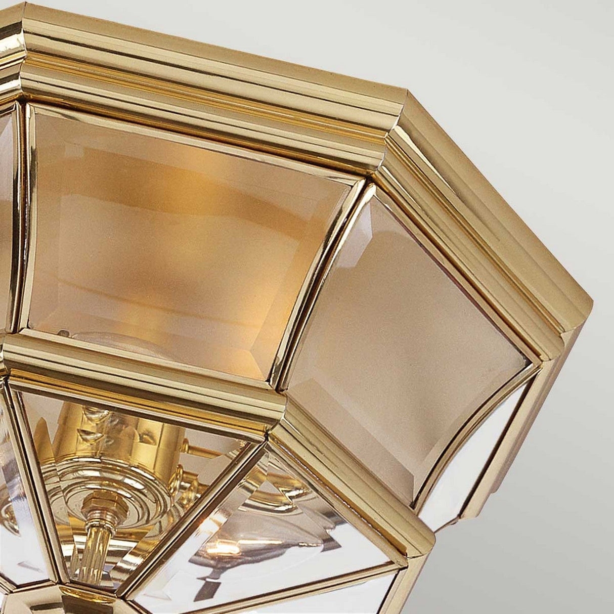 A close-up view of the Newbury Outdoor 3 Light Flush in Lacquered Polished Brass, featuring a gold-framed geometric design with smooth glass panels. The polished brass finish emits a warm glow, accentuating its elegant design and metallic shine.