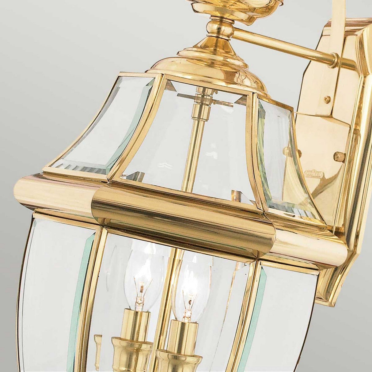 A close-up view of the Newbury Outdoor 2 Light Large Wall Lantern in lacquered polished brass showcases its clear beveled glass panels and two candle-style light bulbs. The lantern boasts a classic, elegant design and is mounted on a golden wall bracket.