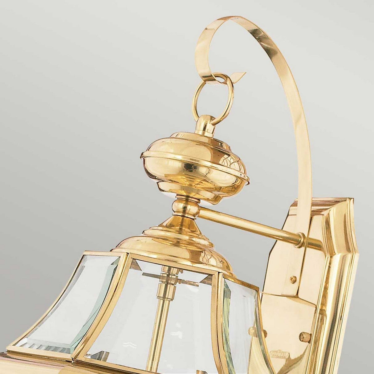A Newbury Outdoor 2 Light Large Wall Lantern in lacquered polished brass, featuring a gold color with a curved arm, clear beveled glass panels, and an elegant design complete with smooth, shiny surfaces and a decorative loop on top is shown against a light gray background.